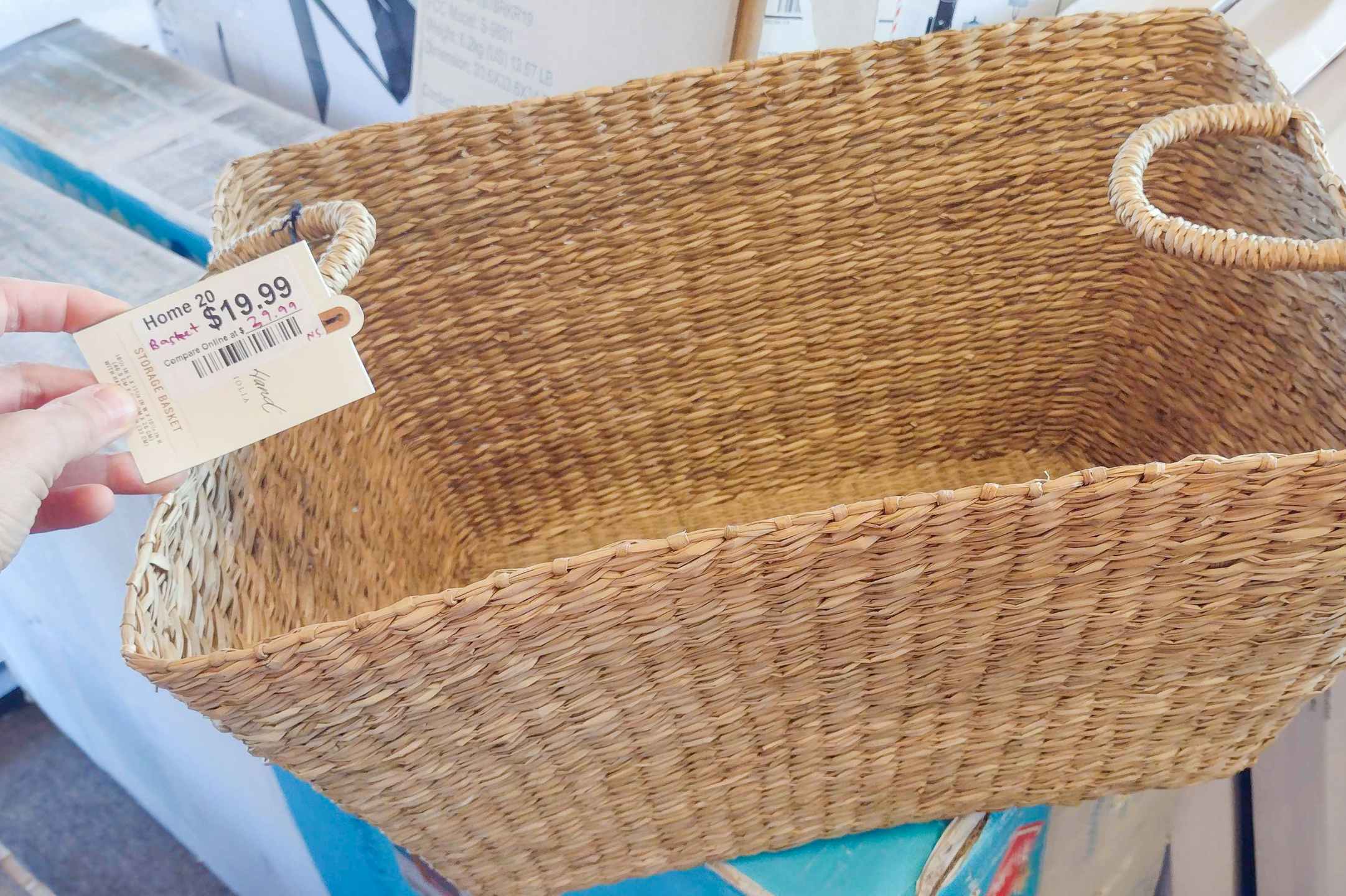 A person's hand holding a tag up for a Hearth and Hand basket sitting on a box.
