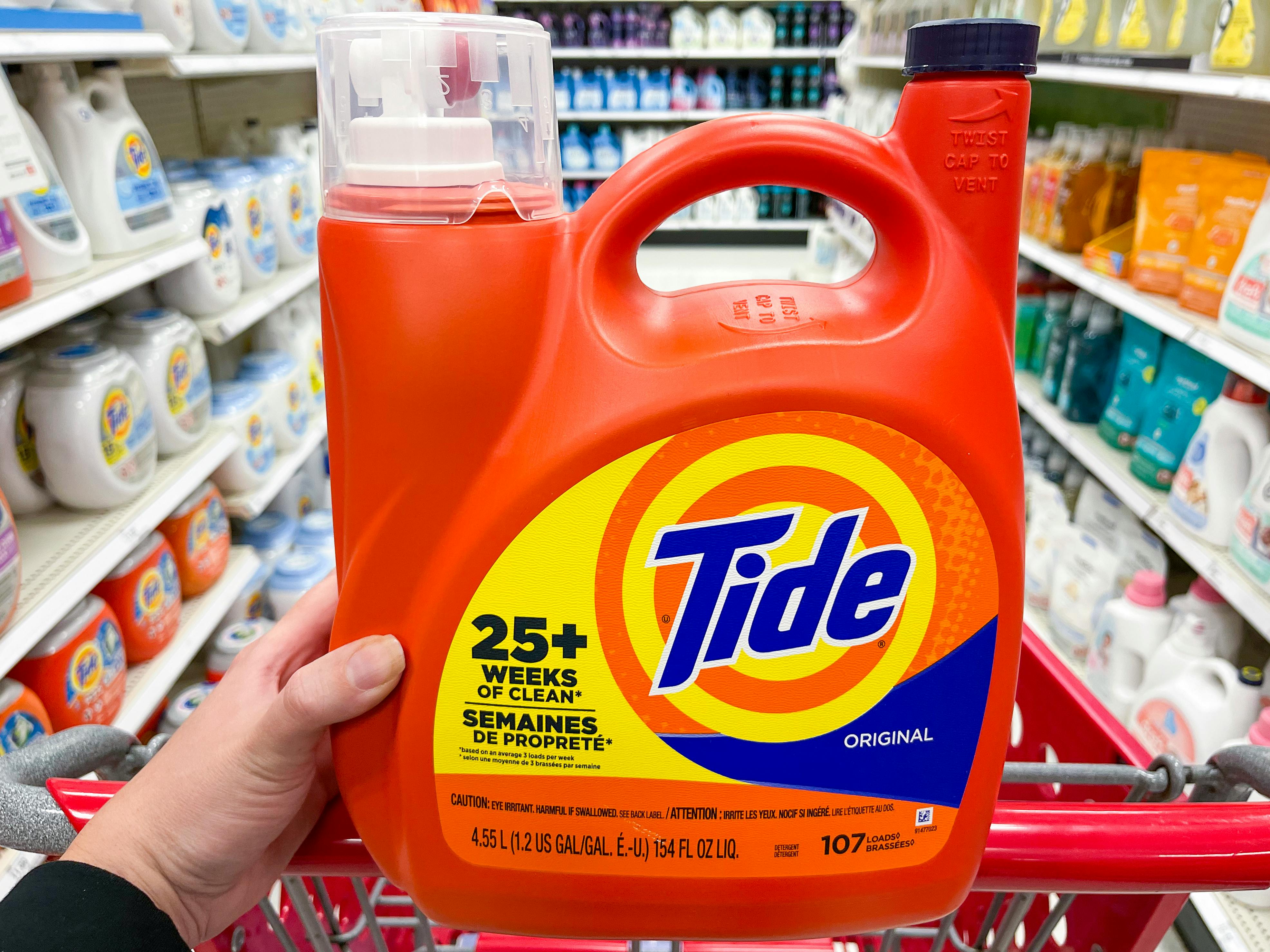 A person's hand resting on the handle of a Target shopping cart holding up a 1.2 gallon container of Tide Original laundry detergent.