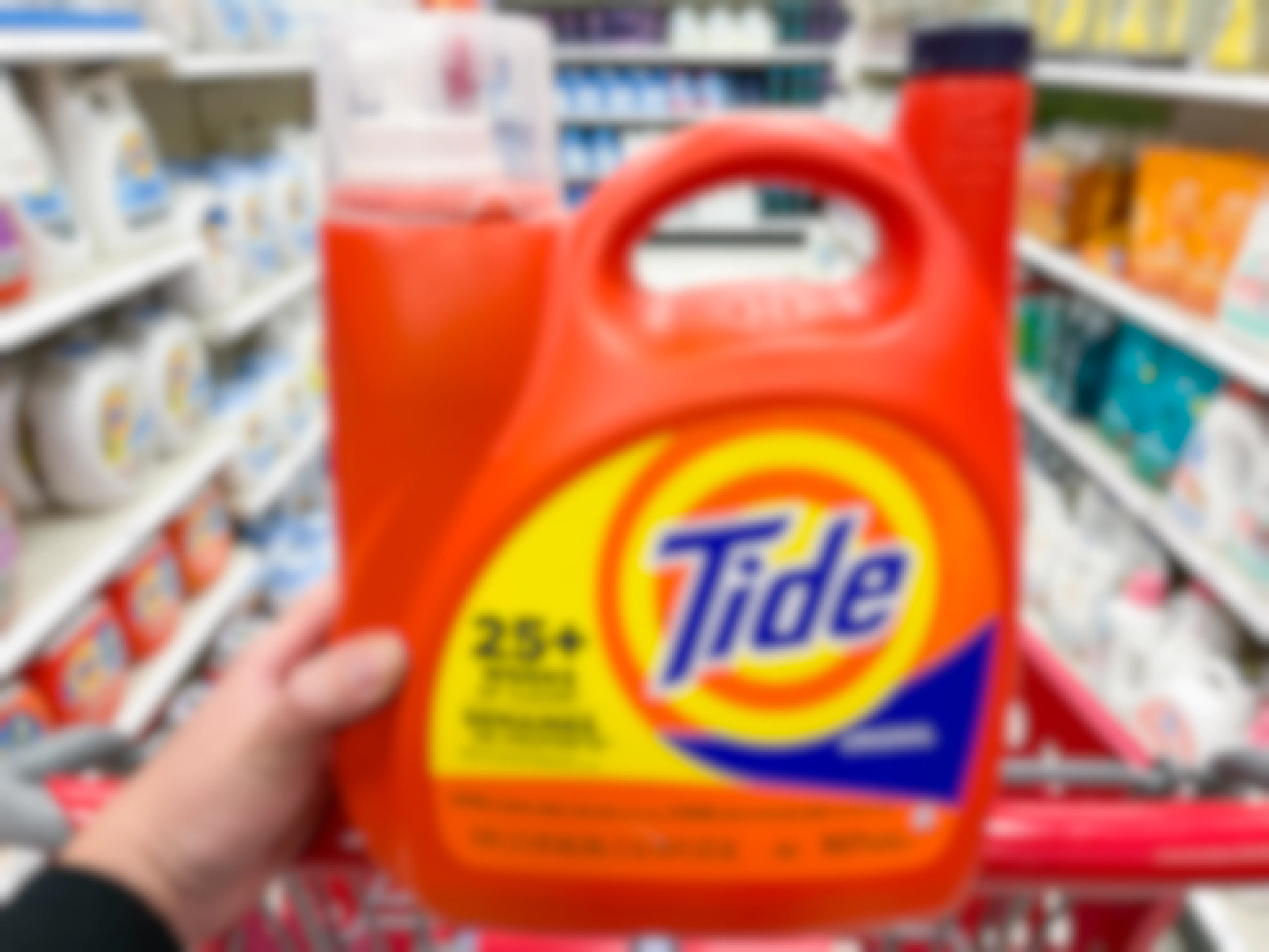 A person's hand resting on the handle of a Target shopping cart holding up a 1.2 gallon container of Tide Original laundry detergent.