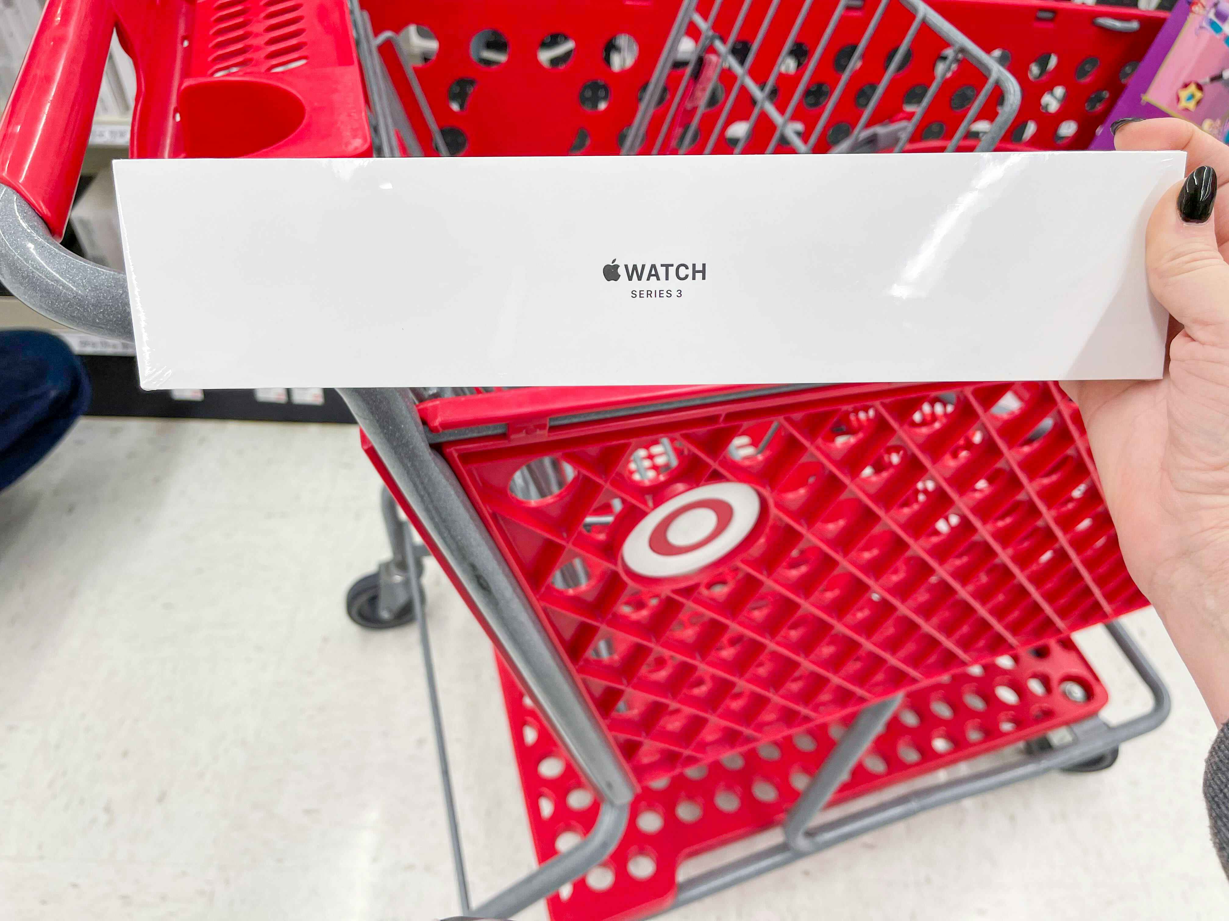 A person's hand holding an Apple Watch Series 3 box in front of a Target shopping cart.