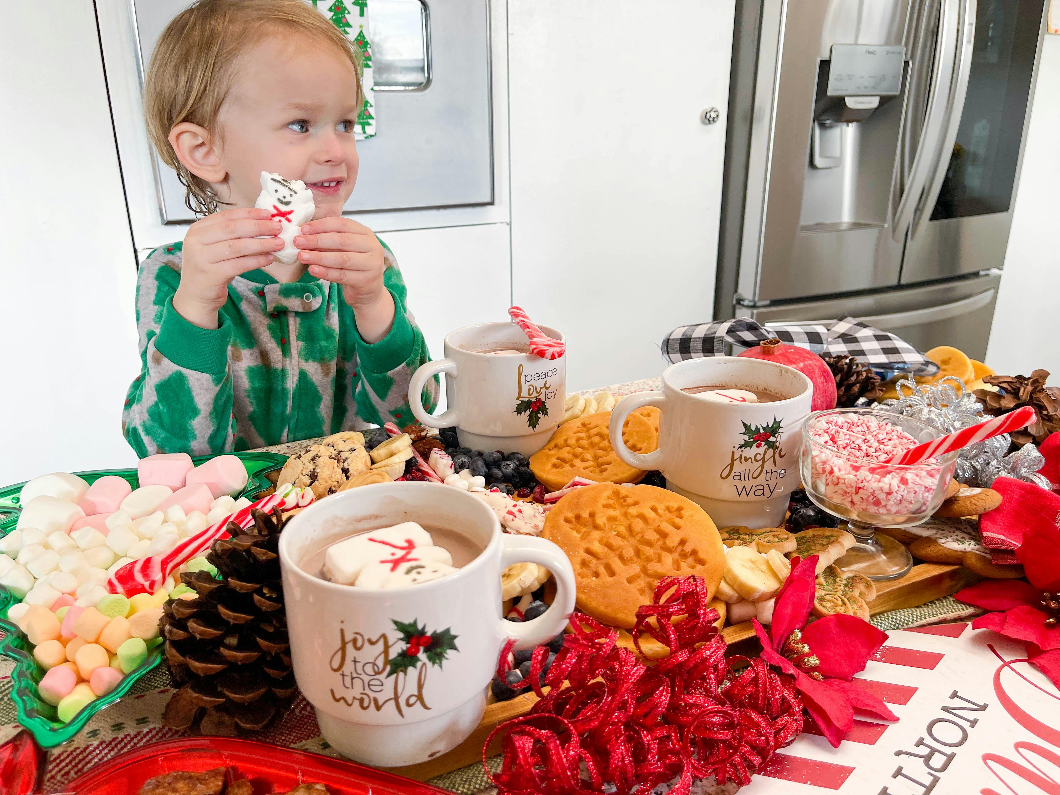 little boy eating off a holiday charcuterie board with hot coco, cookies, and waffles 