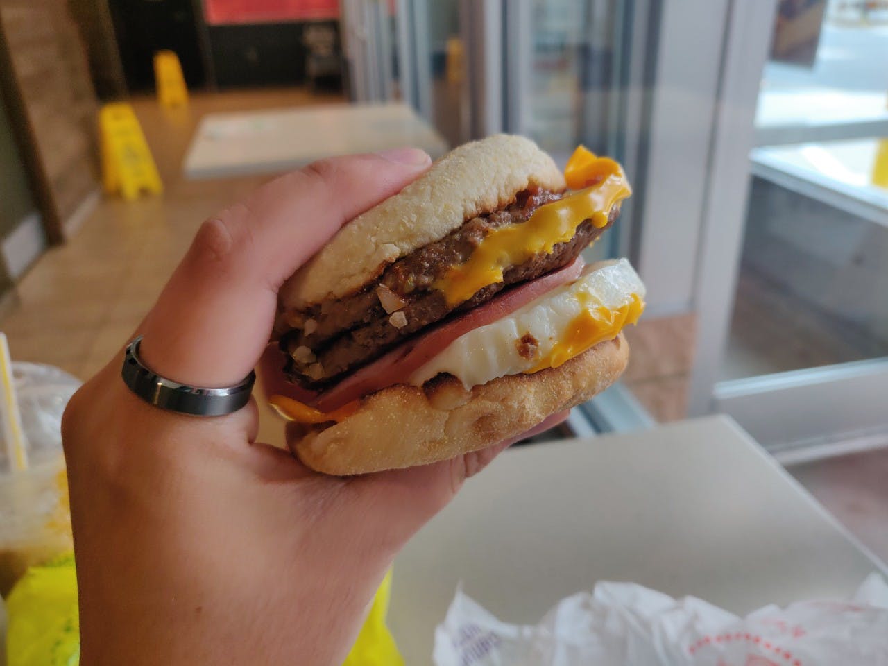 A person's hand holding up a McDonald's breakfast sandwich at a table inside McDonald's.