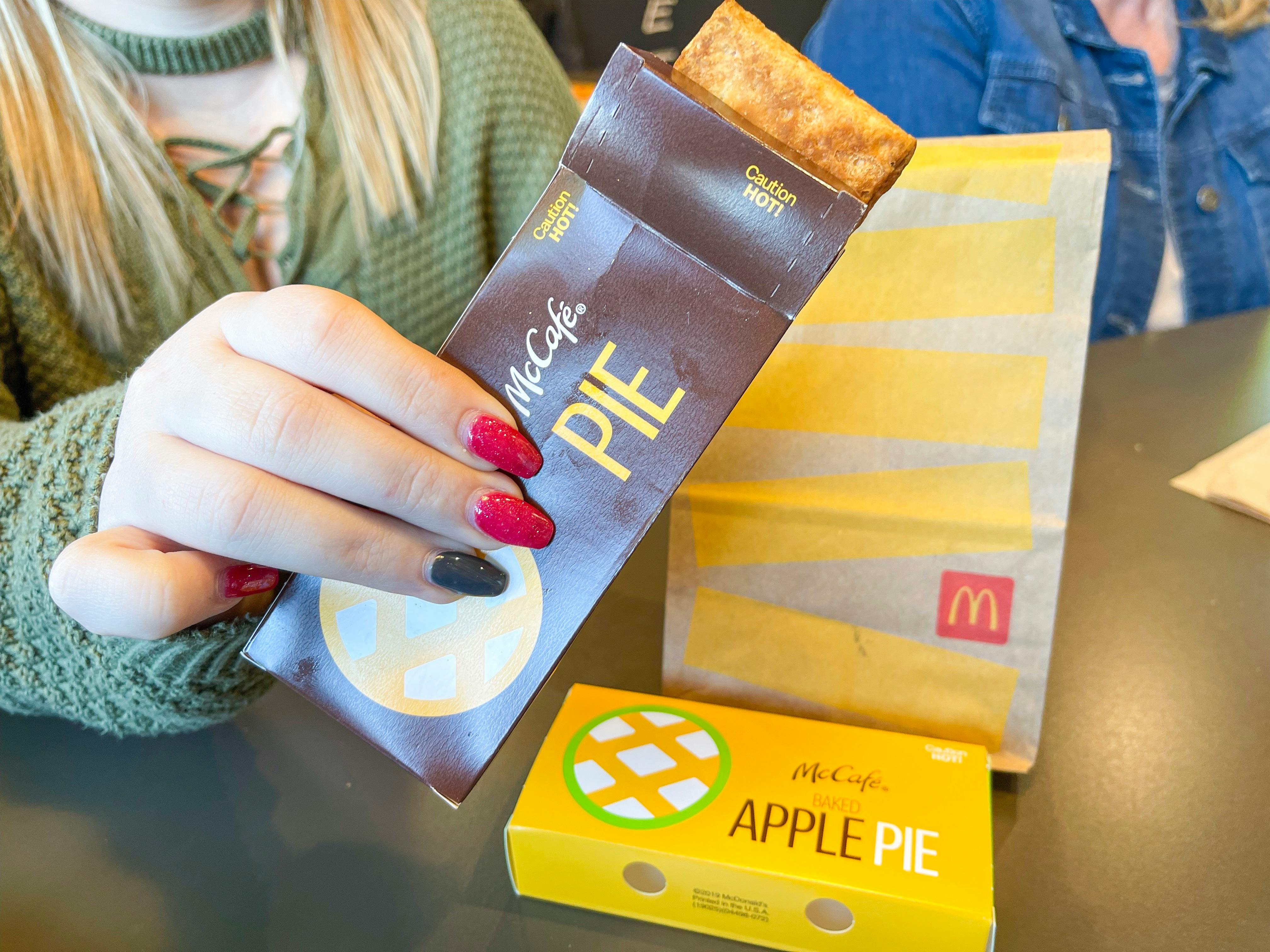 A person holding a McCafe pie in a box in front of a McCafe Apple Pie and McDonald's bag sitting on the table.