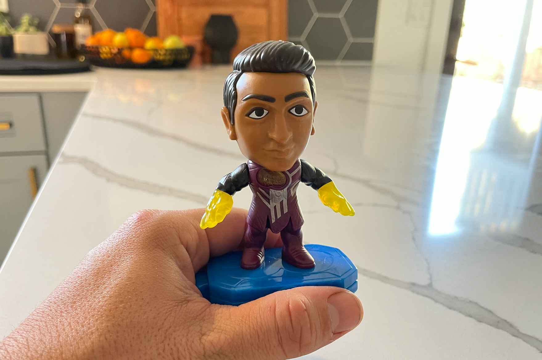 A person's hand holding a McDonald's Happy Meal toy (Kingo from The Eternals) in front of a kitchen counter.