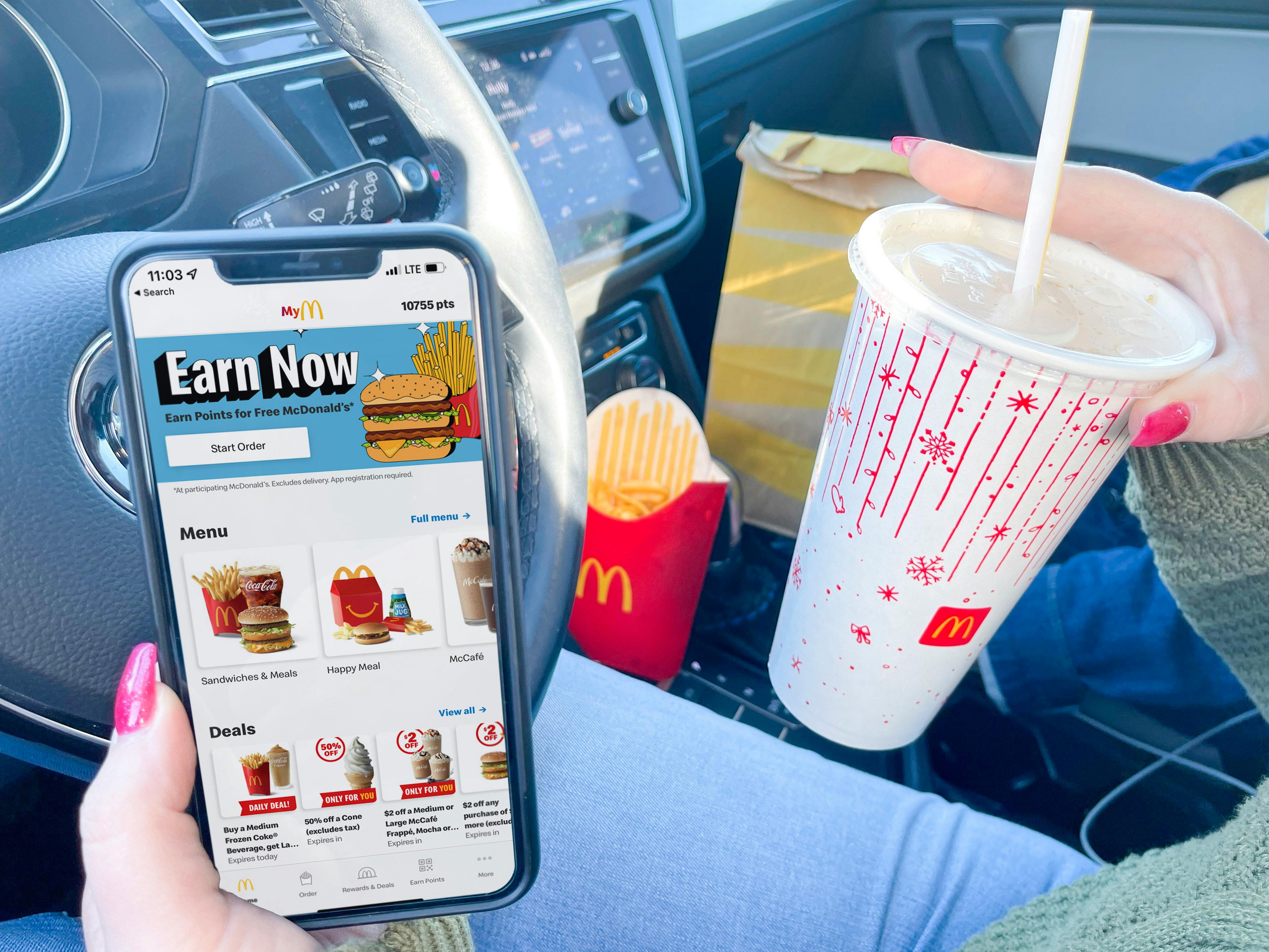 A person sitting in a car while holding an iPhone which is open to the McDonald's mobile app Menu in one hand, and a McDonalds fountain drink in the other hand. On the passenger's seat in the background, there is a box of McDonald's fries and a McDonald's take out bag.