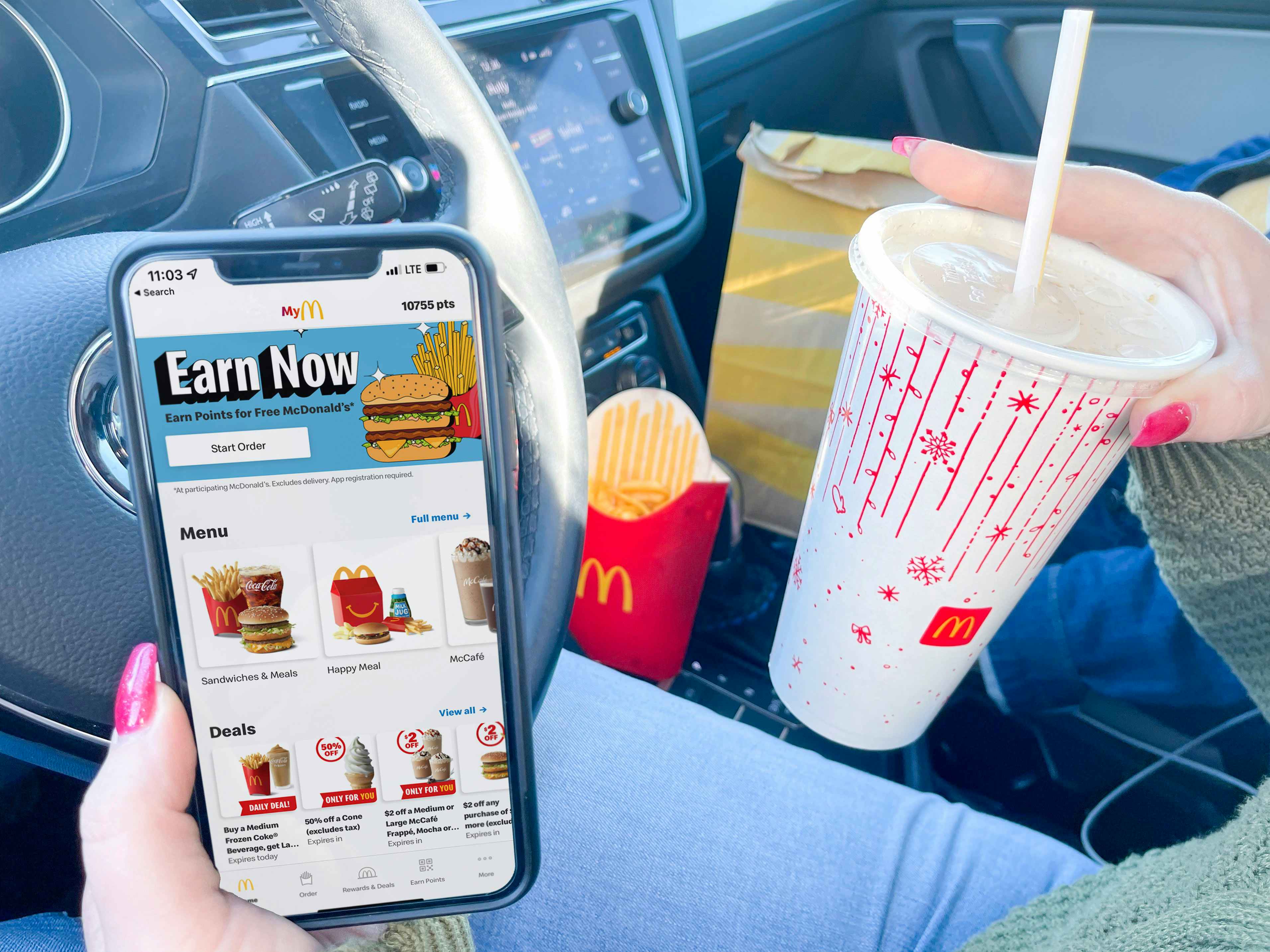 A person sitting in a car holding an iPhone which is open to the McDonald's mobile app Menu in one hand, and a McDonalds fountain drink in the other hand. On the passenger's seat in the background, there is a box of McDonald's fries and a McDonald's take out bag.