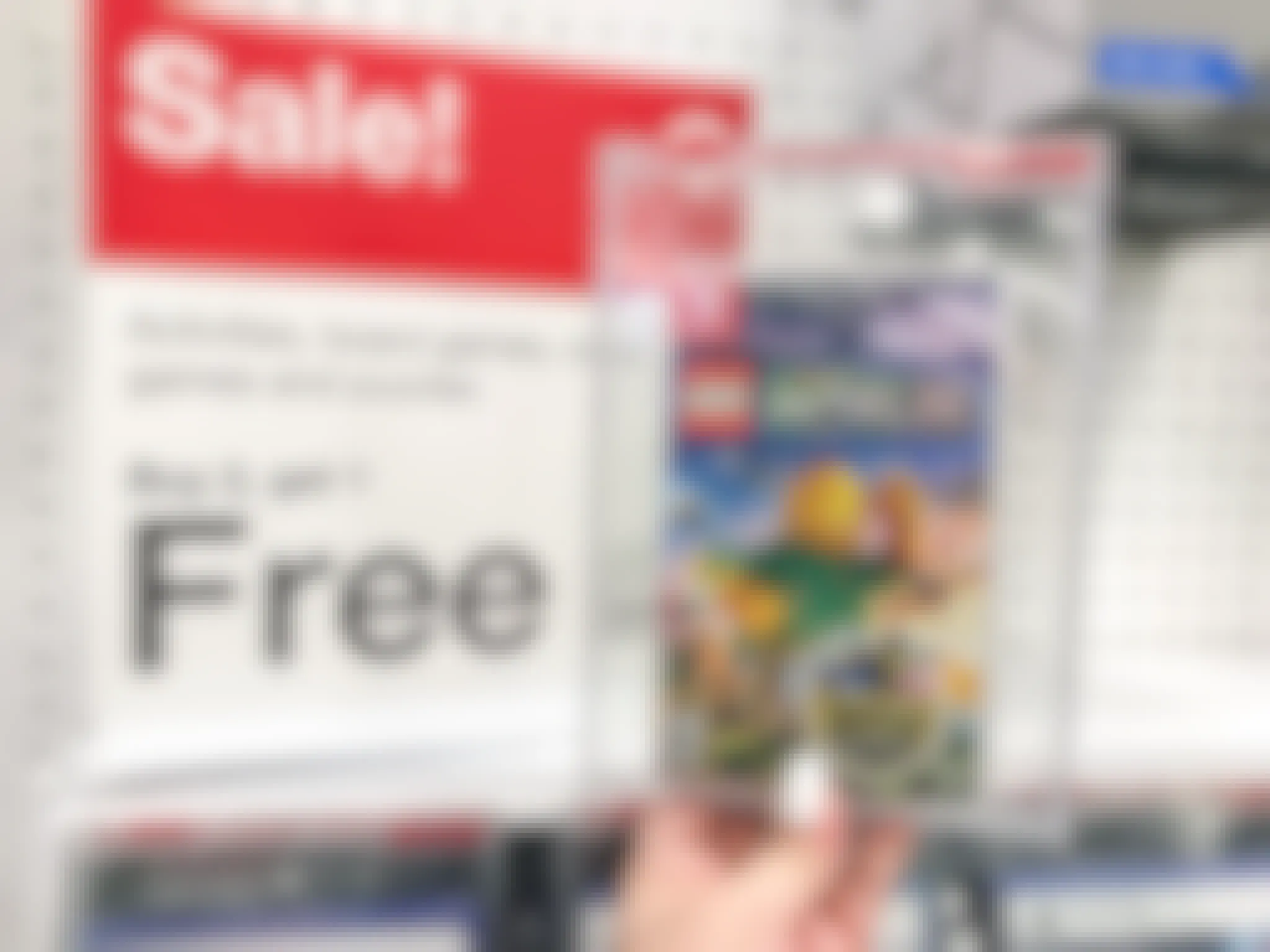 A person's hand holding up a case for LEGO Worlds game for the Nintendo Switch in front of a sign advertising a sale for Buy 2 Get 1 Free on video games and puzzles.