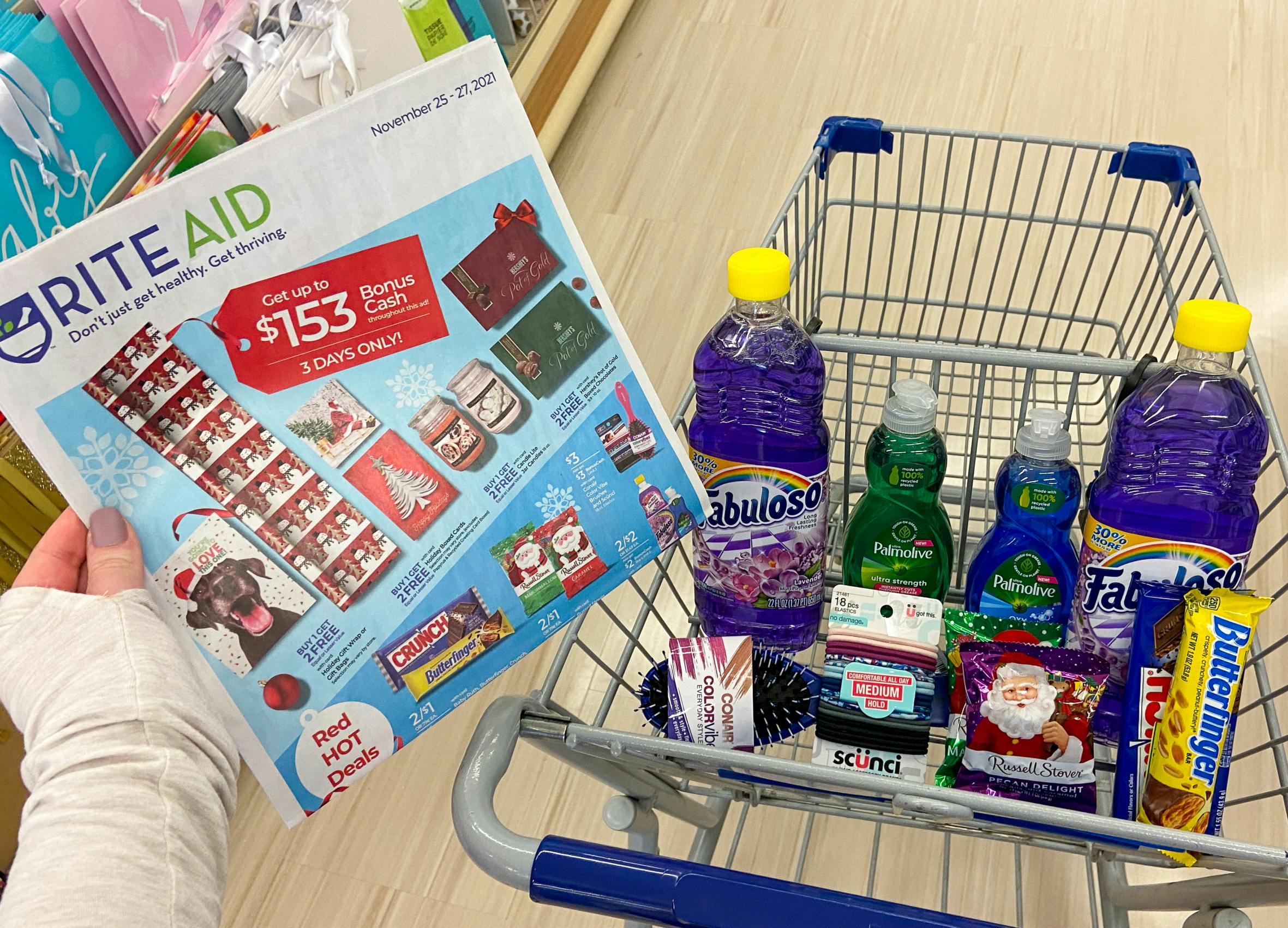 A person's hand holding up the 2021 Rite Aid Black Friday paper ad next to a shopping cart filled with products.