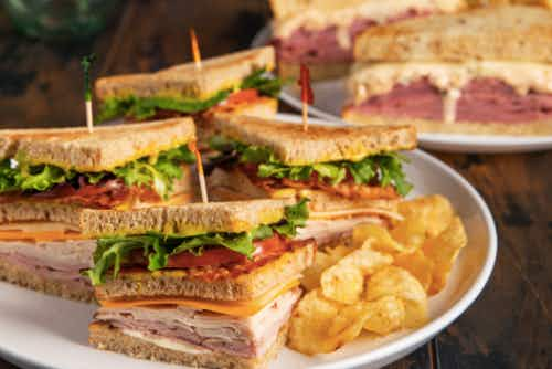 McAlister's ad. Four sandwiches sit atop a white plate with chips beside them. Each has a toothpick with different colors. Two other sandwiches are blurred in the background.