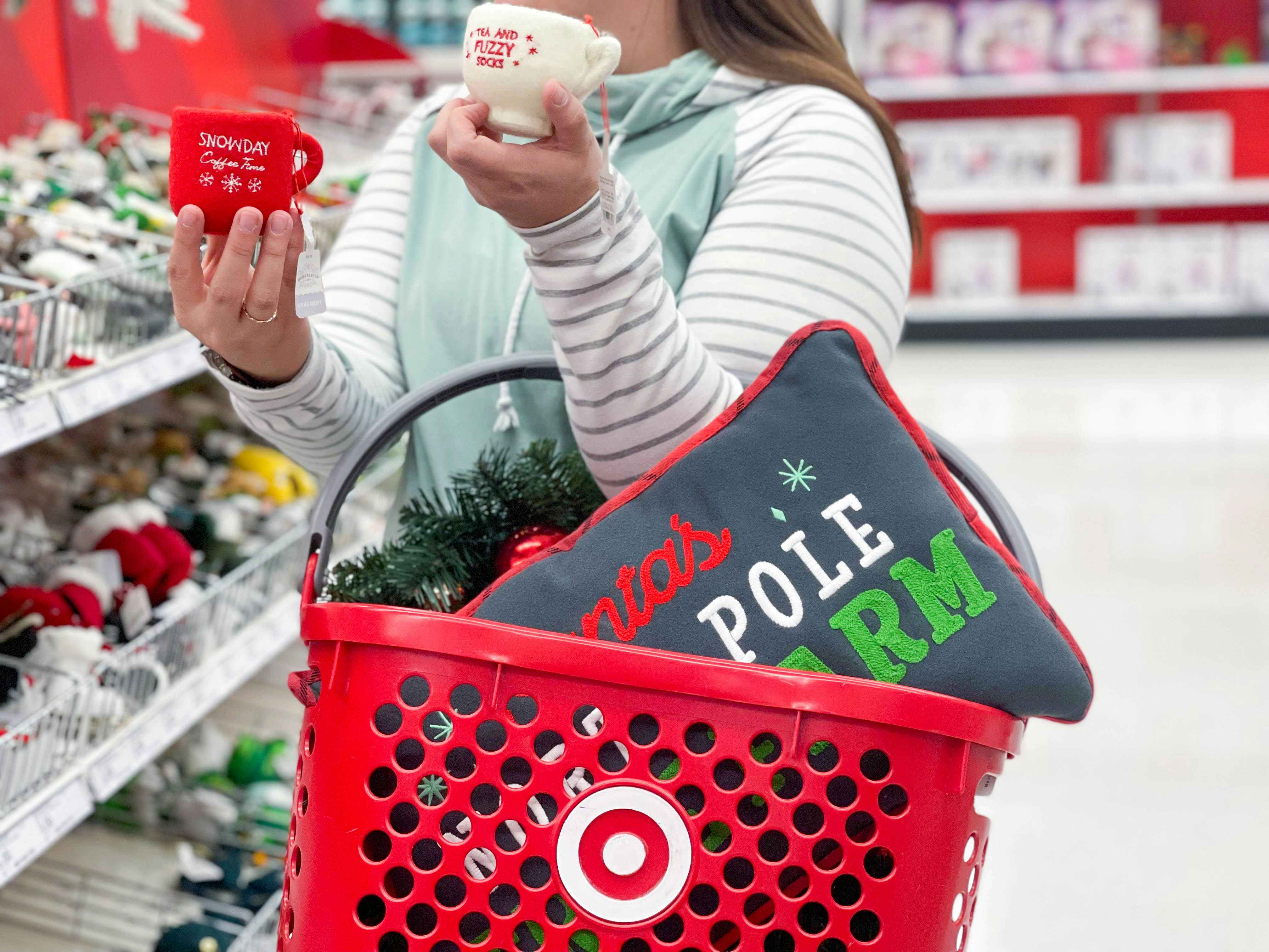 A person carrying a Target hand basket with a holiday-themed pillow in it on her arm, and holding two holiday-themed mugs in an aisle at Target.
