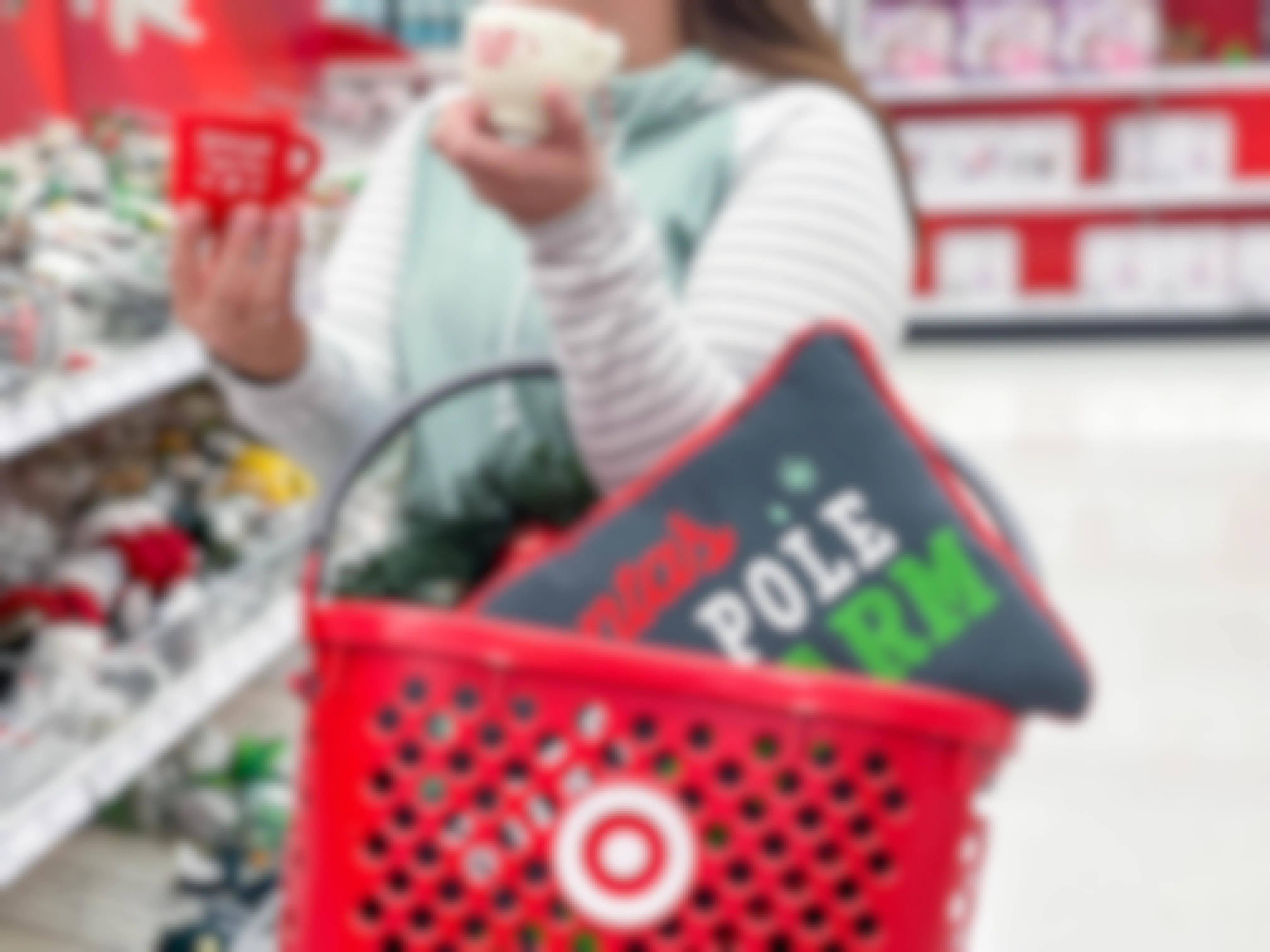 A person carrying a Target hand basket with a holiday-themed pillow in it on her arm, and holding two holiday-themed mugs in an aisle at Target.