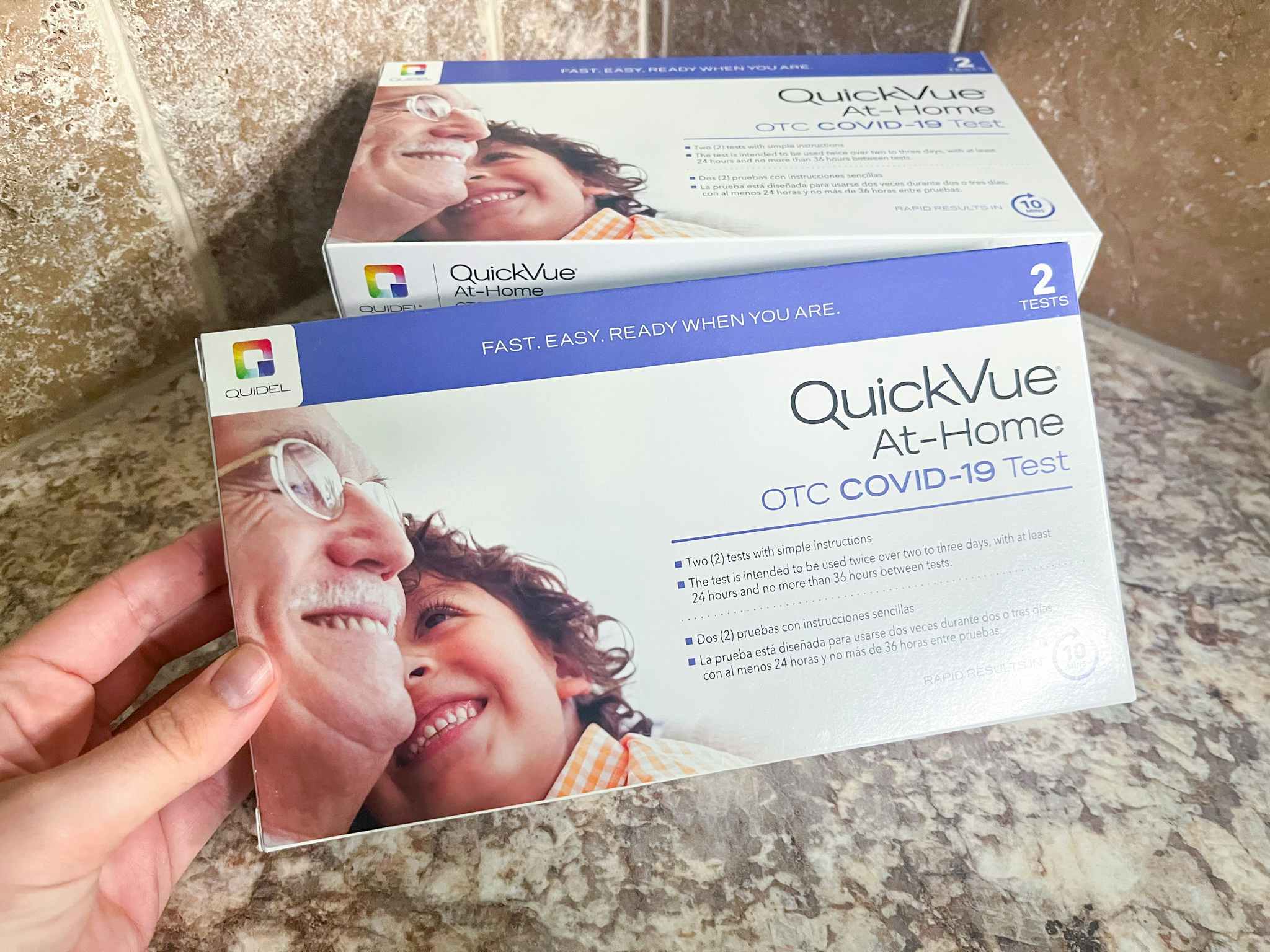 A person's hand holding a QuickVue At-Home COVID-19 test kit box in front of another test kit on a counter.