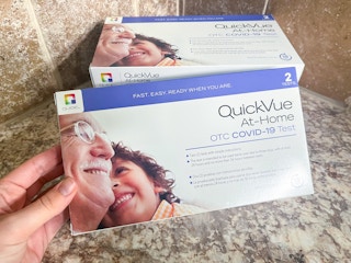 A person's hand holding a QuickVue At-Home COVID-19 test kit box in front of another test kit on a counter.