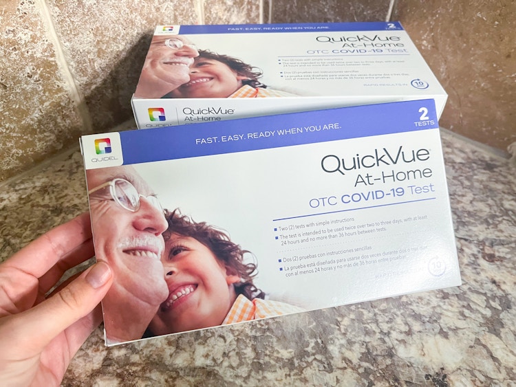 A person's hand holding a QuickVue At-Home COVID-19 test kit box in front of another test kit on a counter.