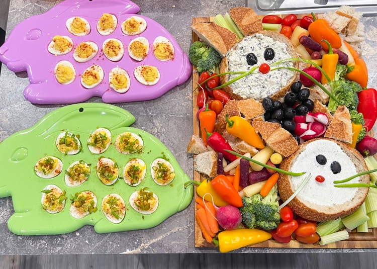 An Easter charcuterie board with bread bunnies and vegetables on a counter next to platters of deviled eggs