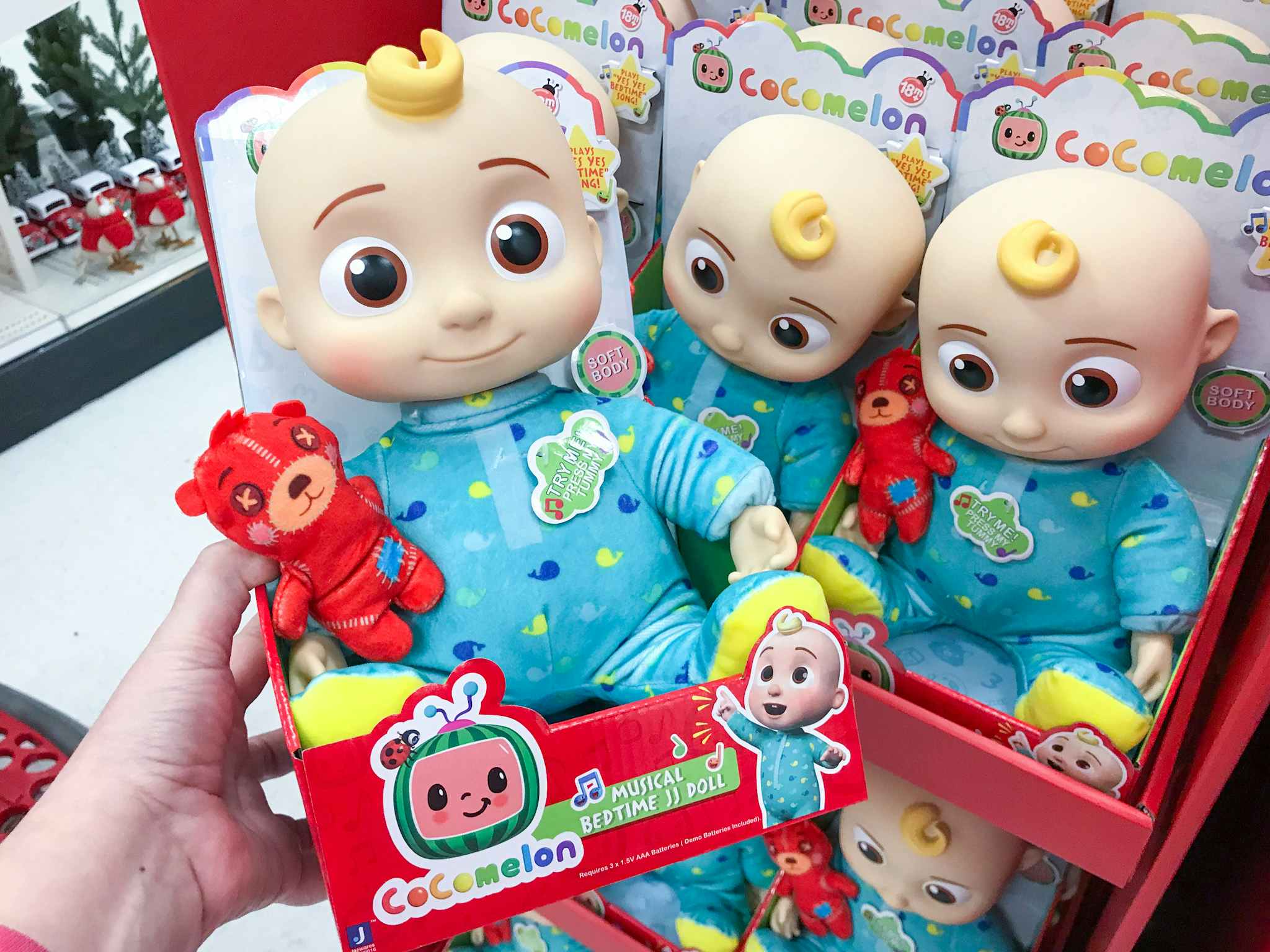 Someone holding a CoComelon Official Bedtime JJ Doll in front of a shelf of more dolls.