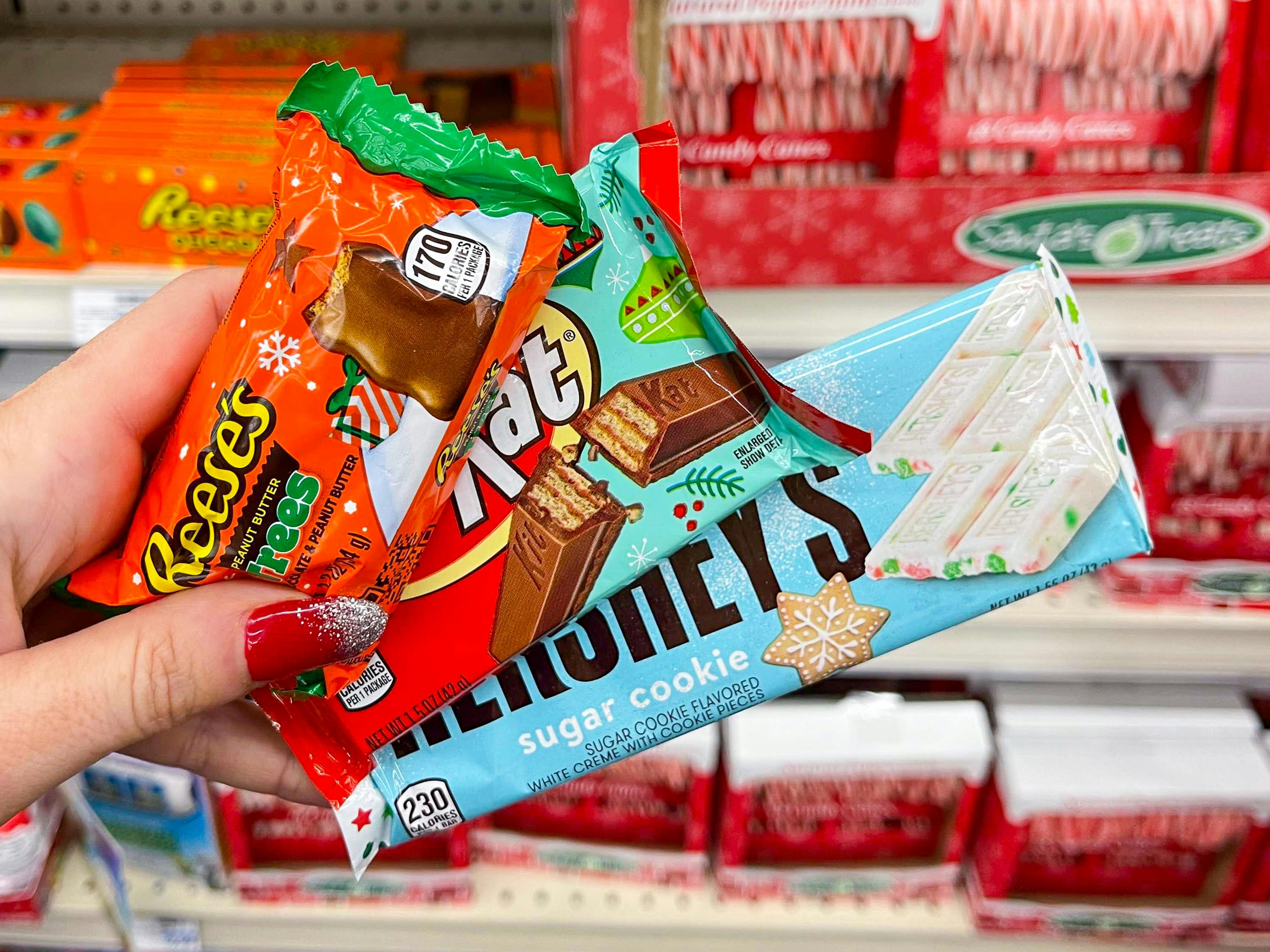 A person's hand holding some holiday candy bars in front of a holiday candy shelf.