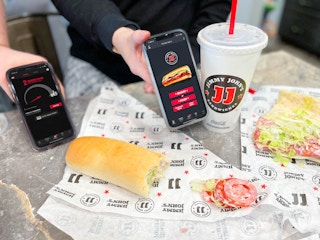 two hands holding phones with jimmy johns app with food on counter