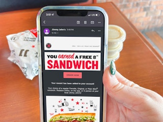A person's hand holding an iPhone displaying an email from Jimmy John's stating the user has earned a free 8" sandwich. A Jimmy John's sandwich wrapped in paper and extra sauces can be seen on the table behind it.