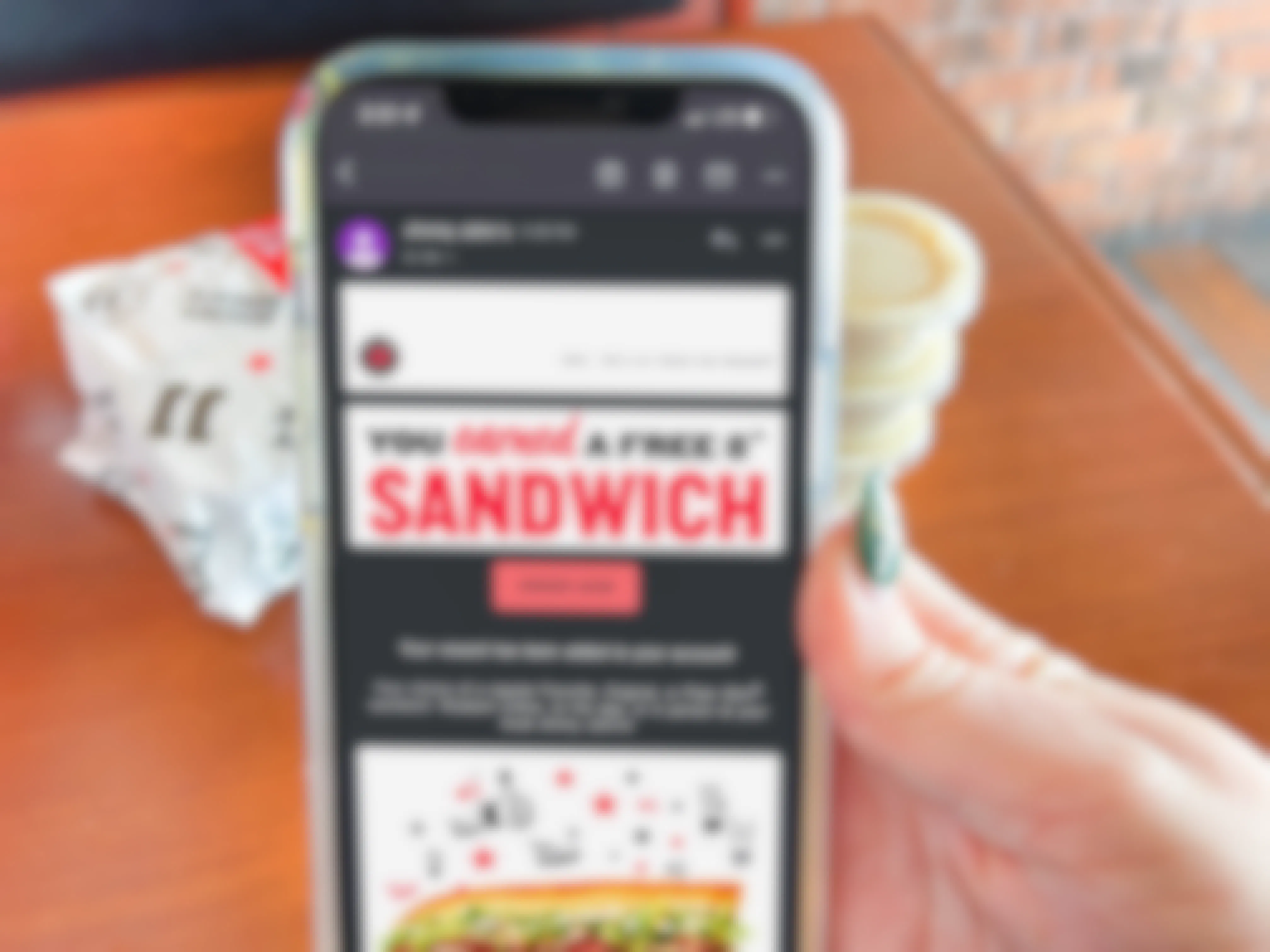 A person's hand holding an iPhone displaying an email from Jimmy John's stating the user has earned a free 8" sandwich. A Jimmy John's sandwich wrapped in paper and extra sauces can be seen on the table behind it.