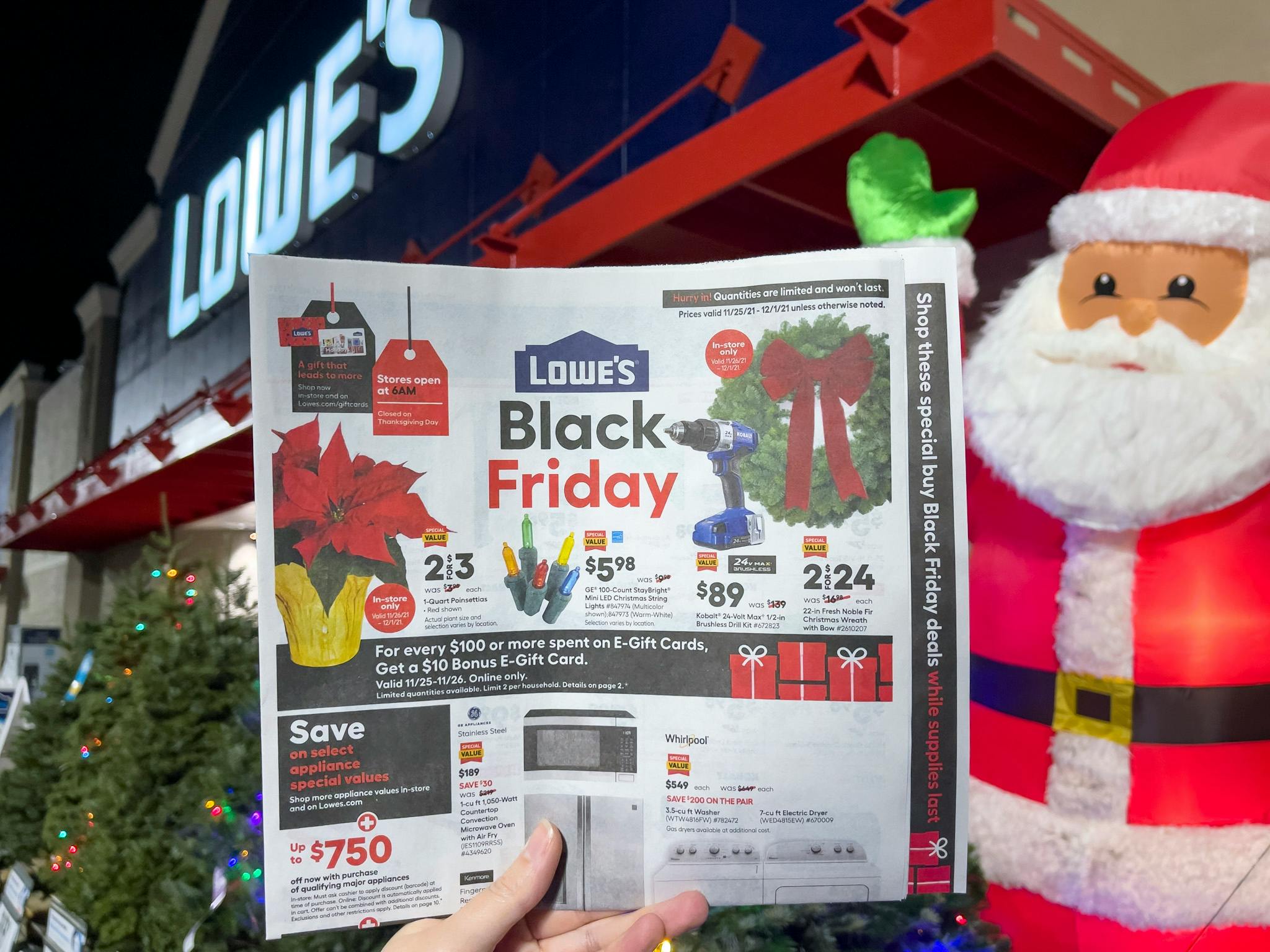 A person's hand holding up a Lowe's Black Friday ad in front of a Lowe's store and a Santa decoration.