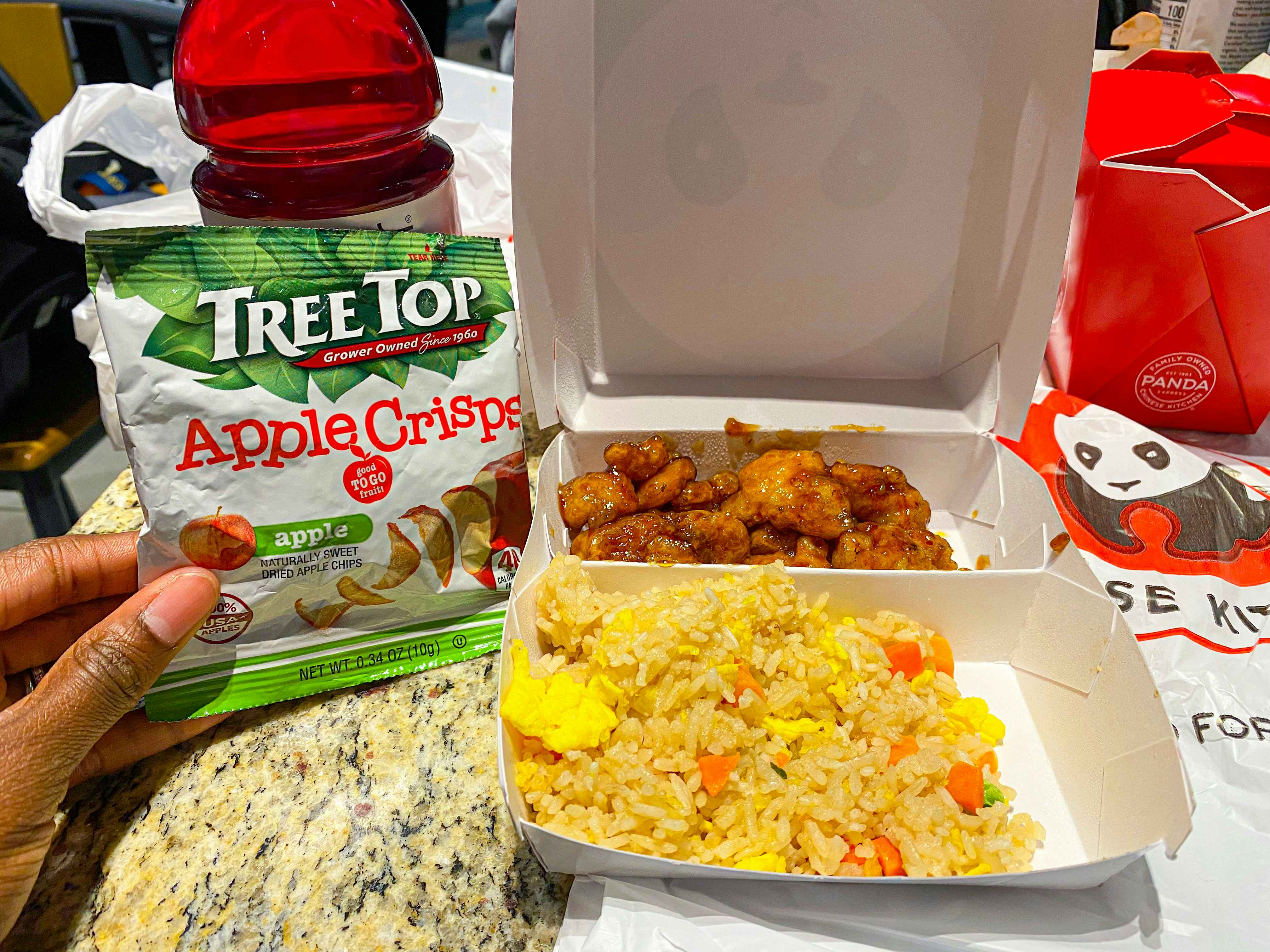 A person's hand holding Tree Top apple crisps next to a Panda Express kids meal including fried rice and orange chicken. 