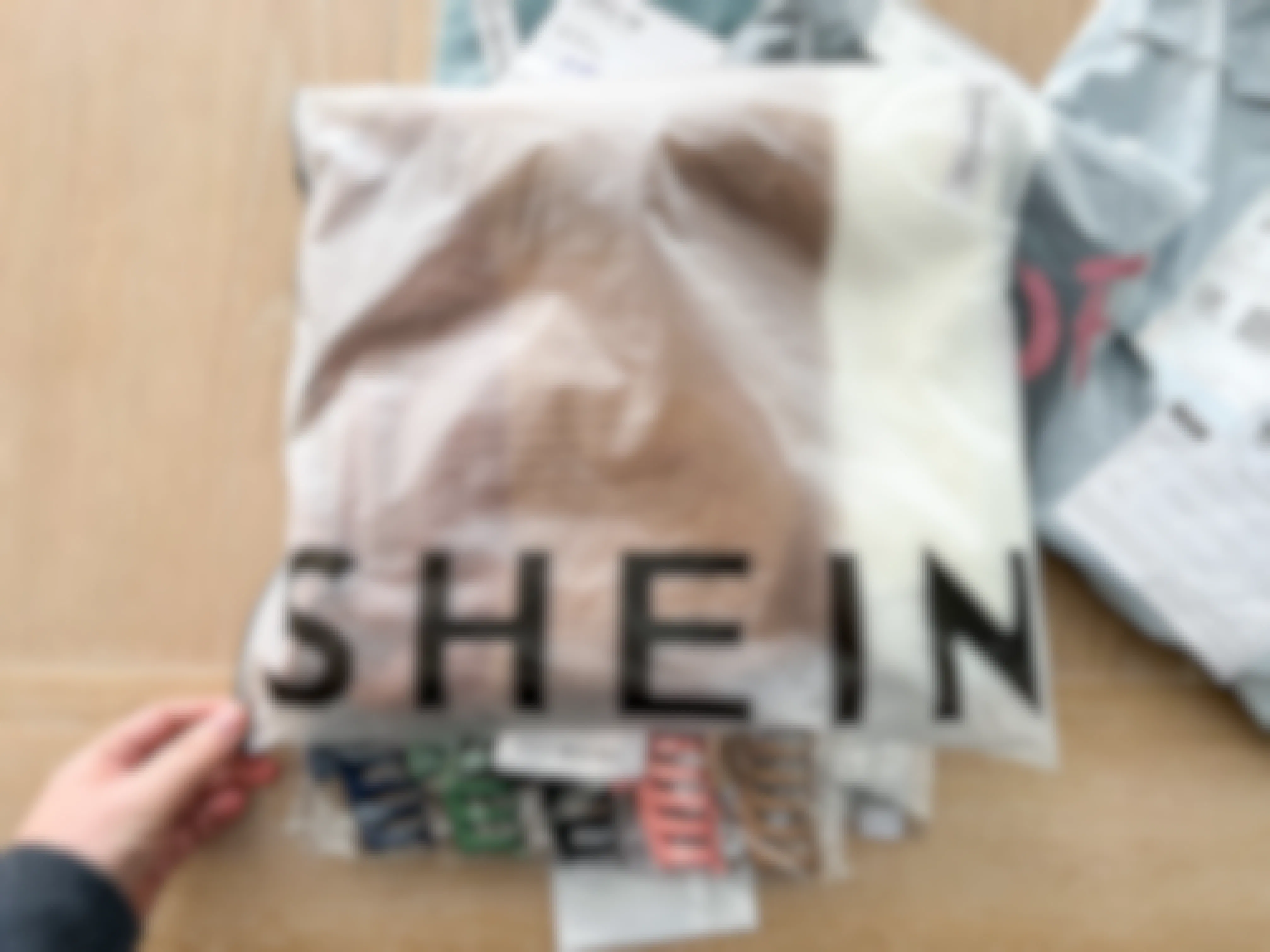 A person's hand grabbing a package of SHEIN clothes sitting on a wooden table.