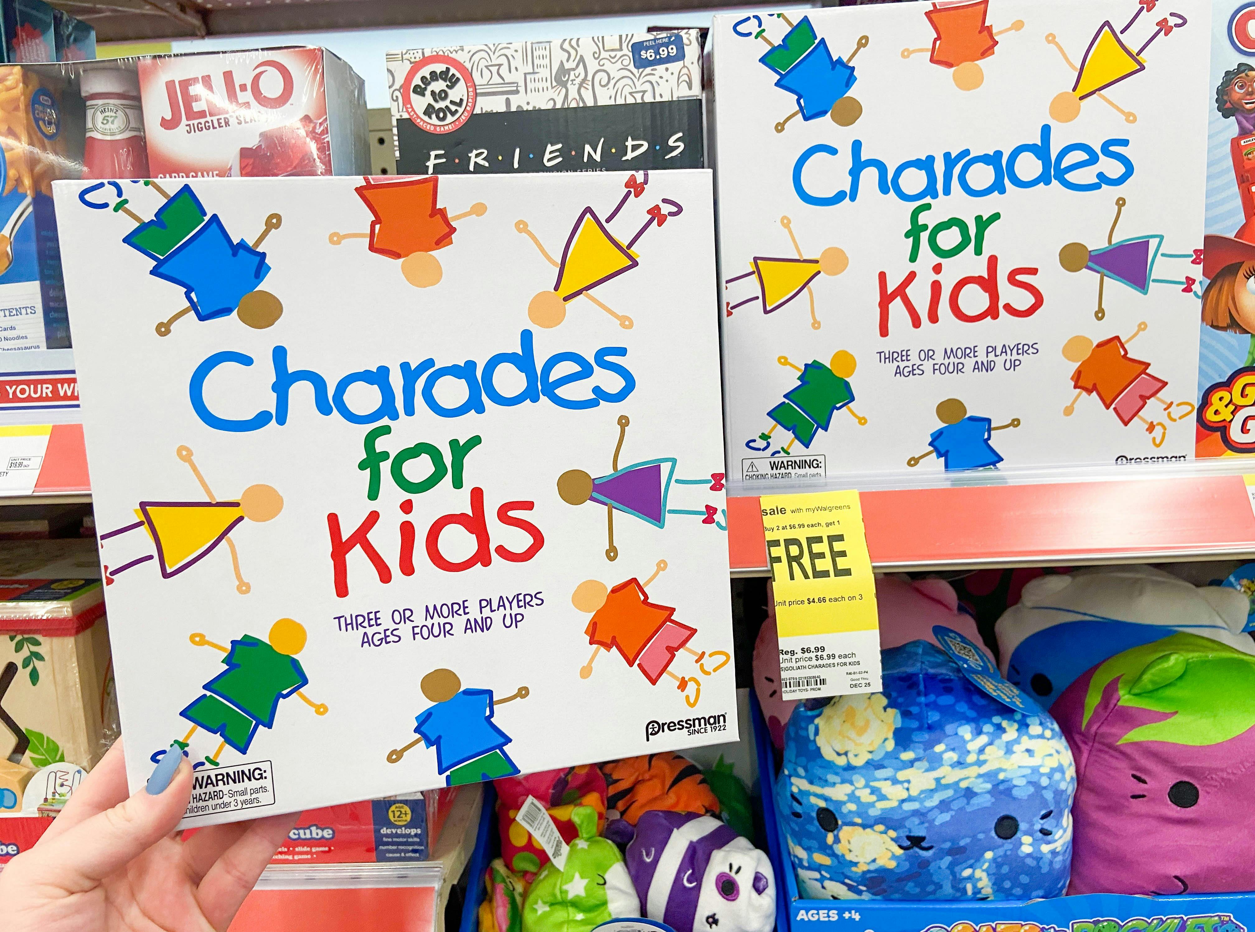A person's hand holding up a board game called Charades for Kids in front of a shelf of more games in Walgreens.