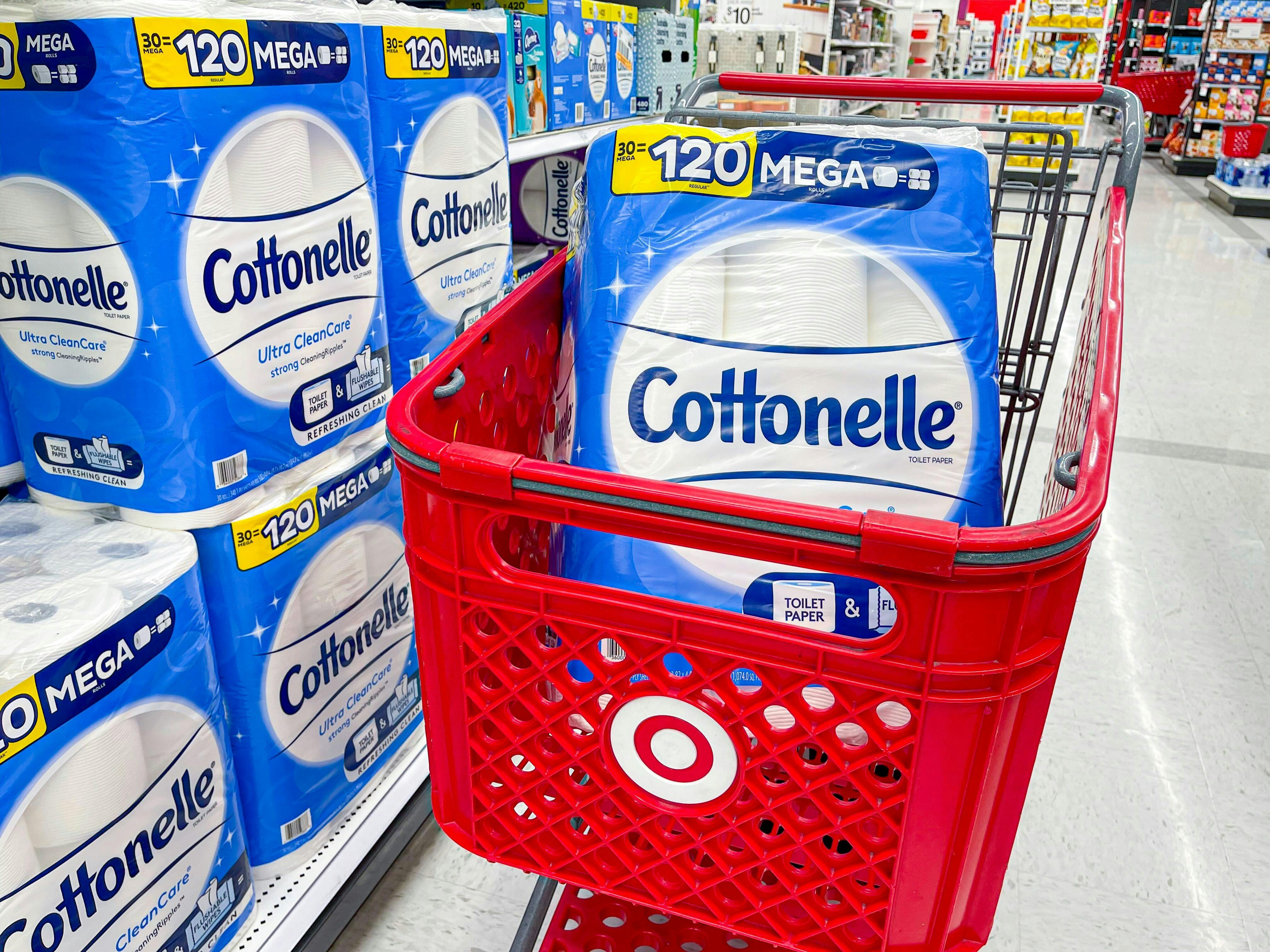 One pack of Cottonelle Mega toilet paper sitting in a Target shopping cart parked in an aisle next to a shelf displaying more Cottonelle toilet paper packs.