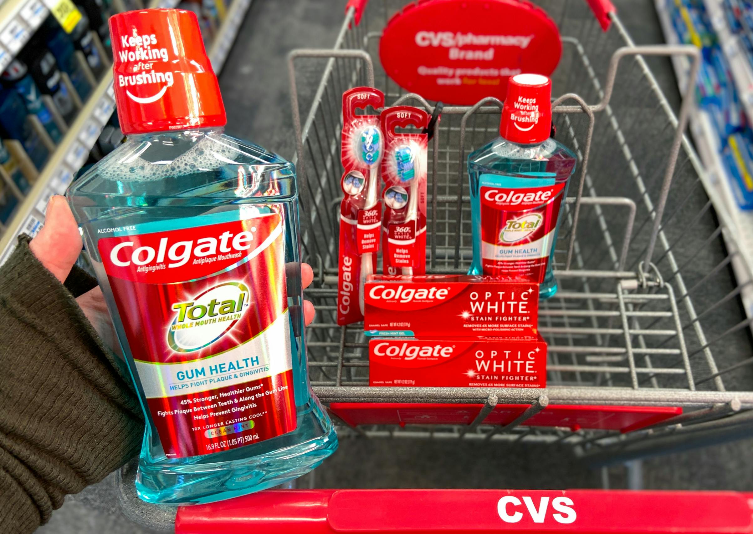 A person's hand holding a bottle of Colgate Total mouthwash above a CVS shopping cart with more Colgate mouthwash, Colgate Optic White toothpaste, and Colgate toothbrushes in the basket.