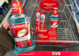 A person's hand holding a bottle of Colgate Total mouthwash above a CVS shopping cart with more Colgate mouthwash, Colgate Optic White toothpaste, and Colgate toothbrushes in the basket.