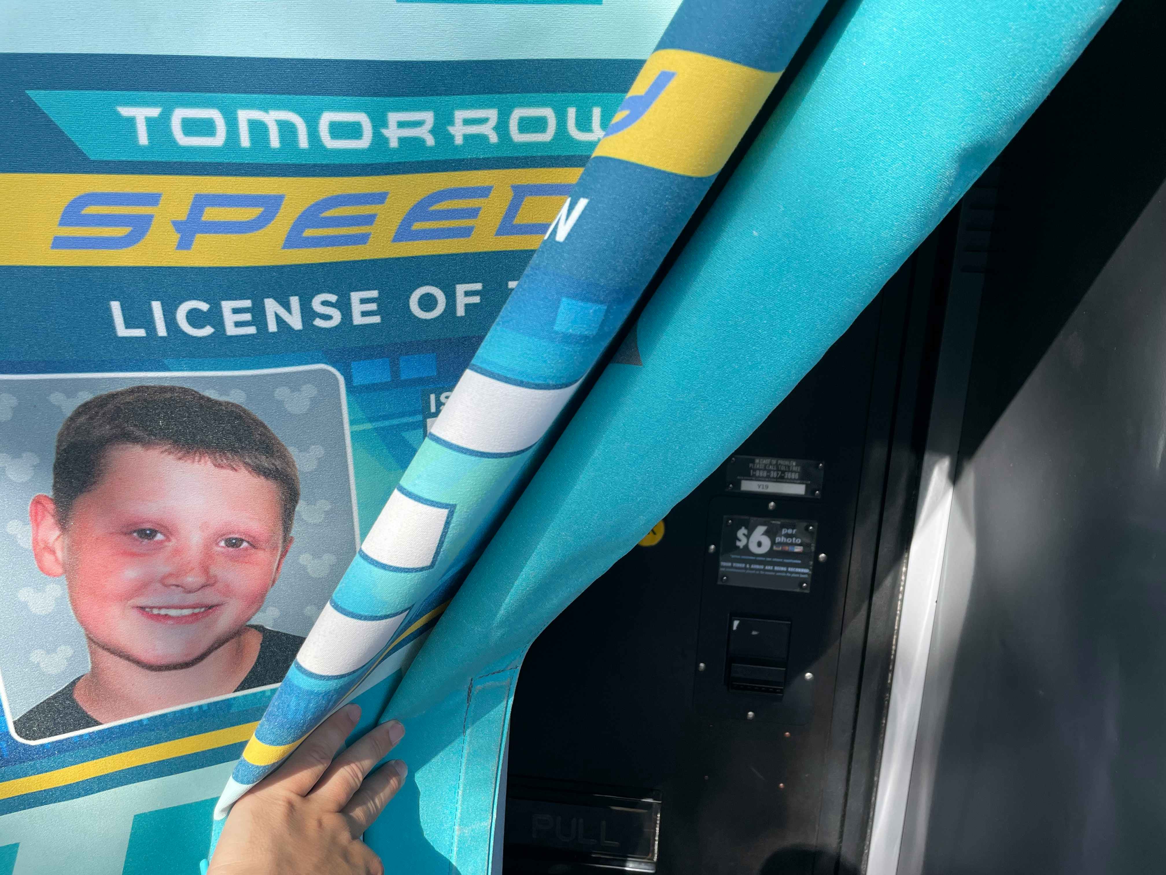 A person's hand opening the curtain to the Tomorrowland Speedway driver's license booth, revealing the $6 price tag.