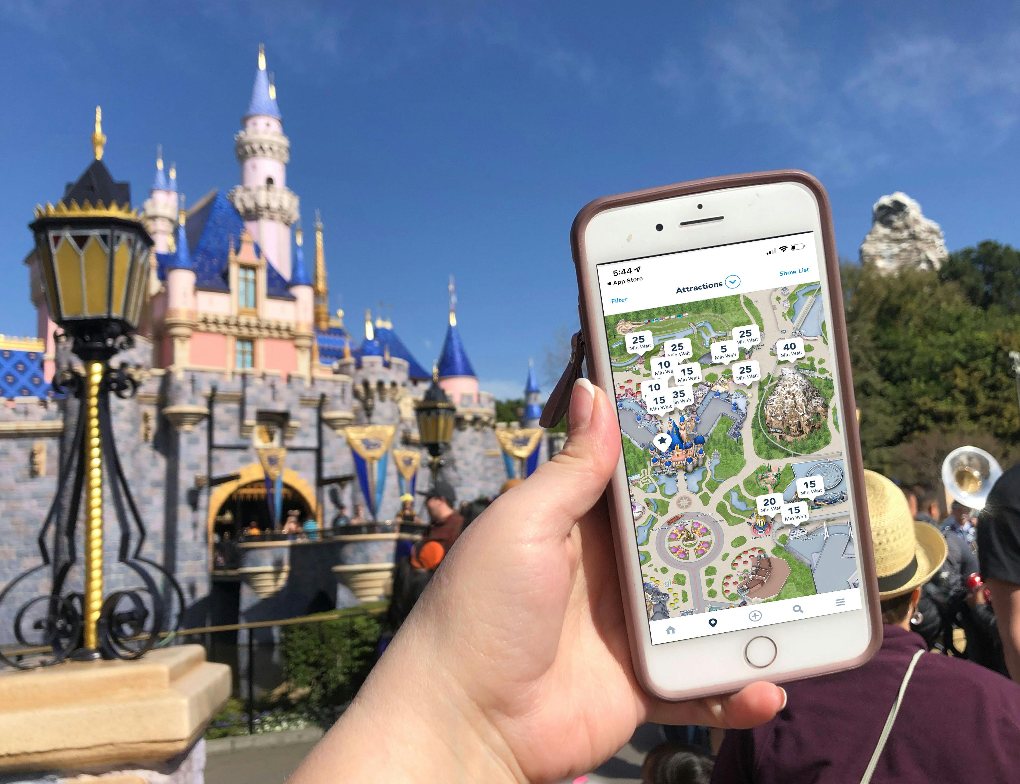 A person's hand holding up a phone displaying the Disneyland app with wait times on a map in front of the Magic Kingdom castle.