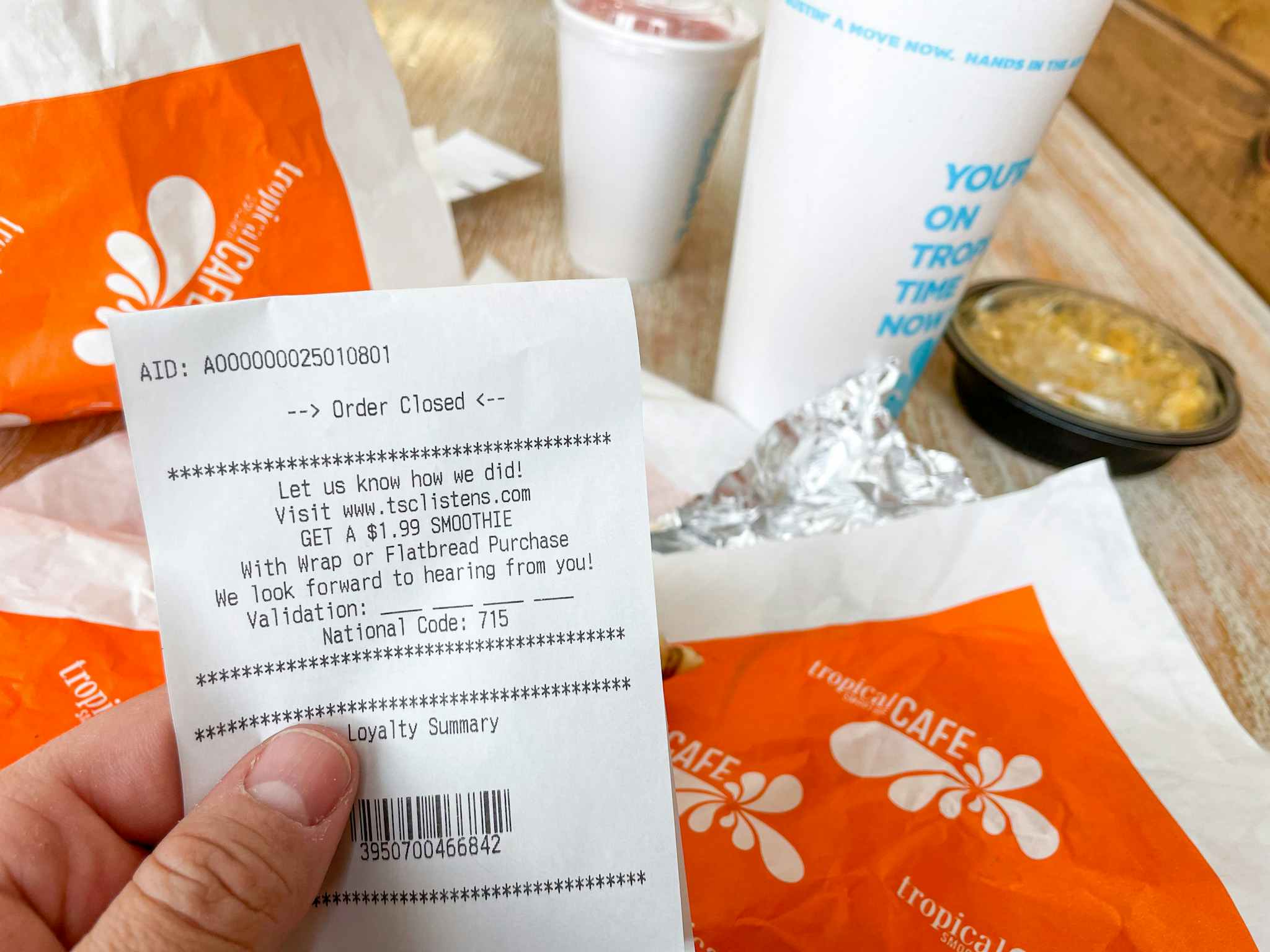 A person's hand holding a receipt from Tropical Smoothie Cafe in front of their smoothie and food on the table.