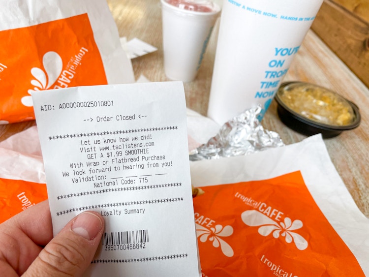 A person's hand holding a receipt from Tropical Smoothie Cafe in front of their smoothie and food on the table.