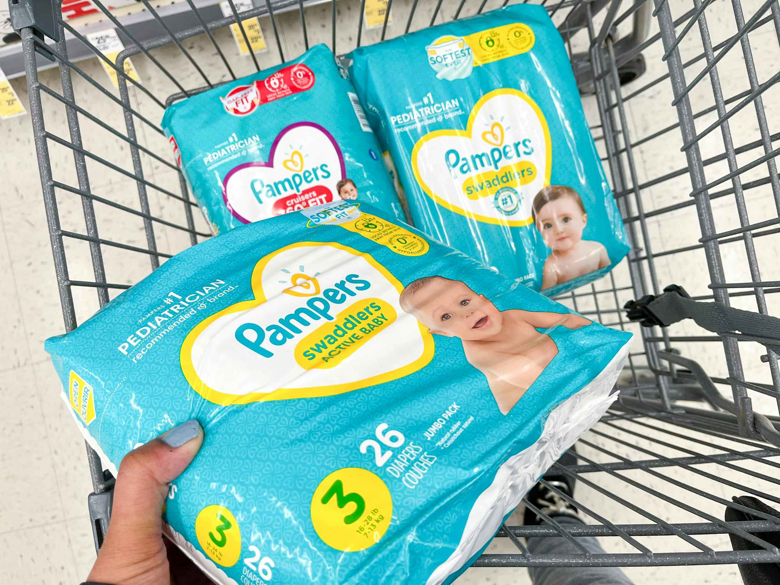 A person putting a pack of Pameprs diapers in a grocery cart with other diapers in it.