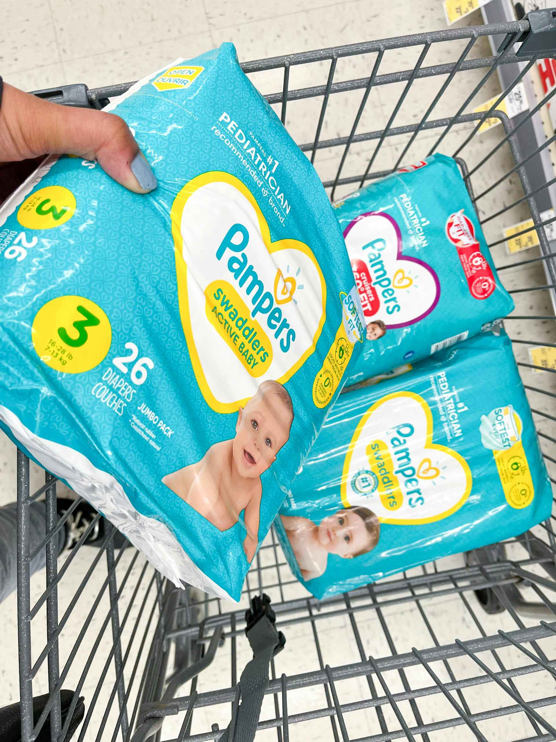 A person putting a pack of Pameprs diapers in a grocery cart with other diapers in it.
