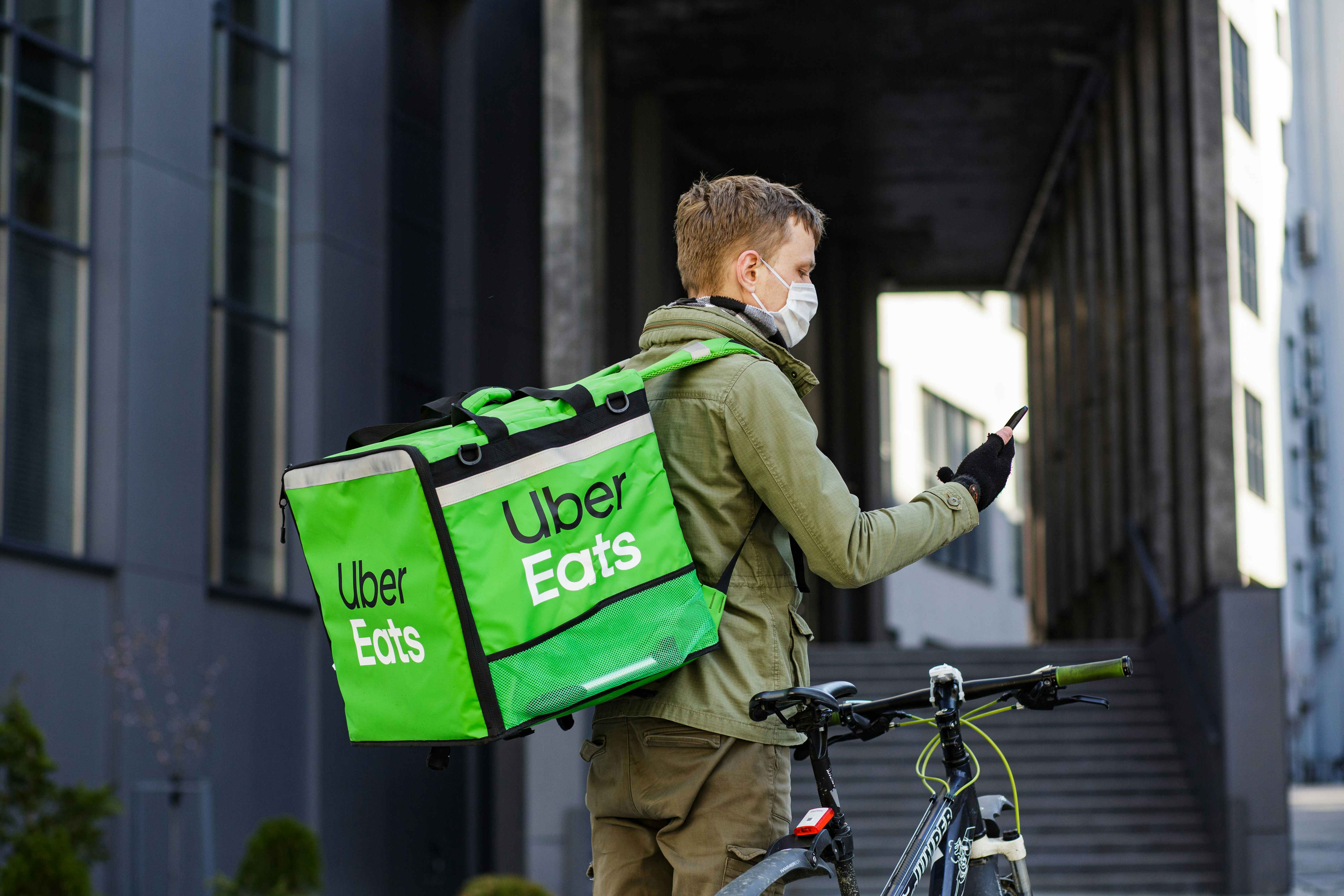A masked Uber Eats delivery person with a cooler backpack parking their bike to deliver an order.