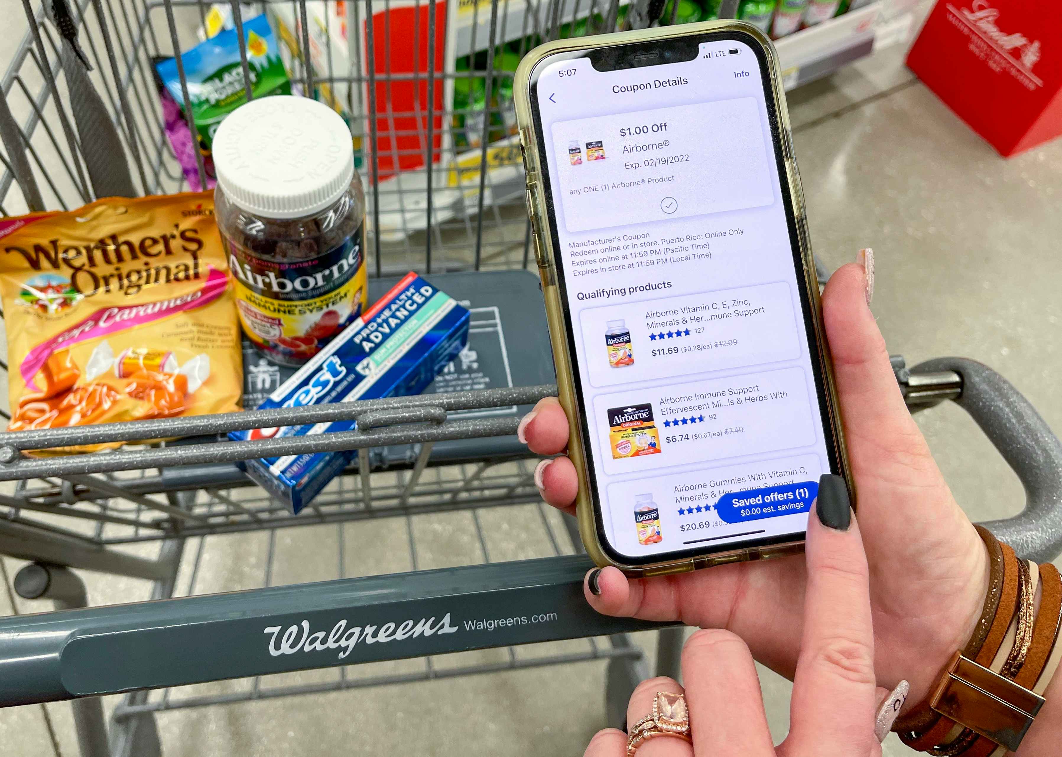 someone holding phone with walgreens app next to cart with candy, vitamins, and toothpaste
