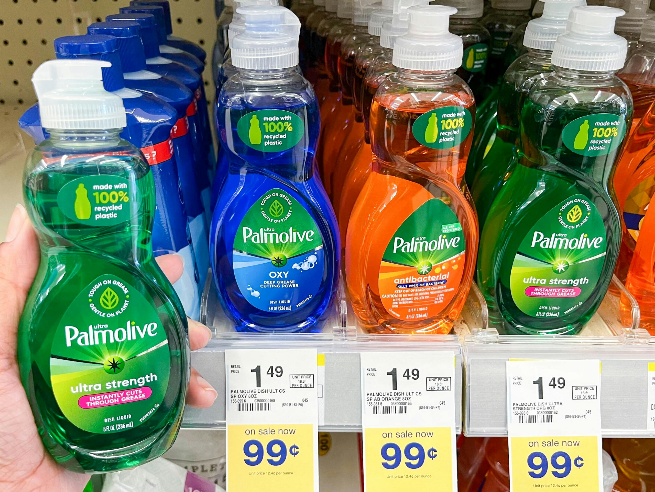 A person's hand holding a bottle of Palmolive Ultra Strength liquid dish soap next to a shelf of different variations of Palmolive liquid dish soap at Walgreens.