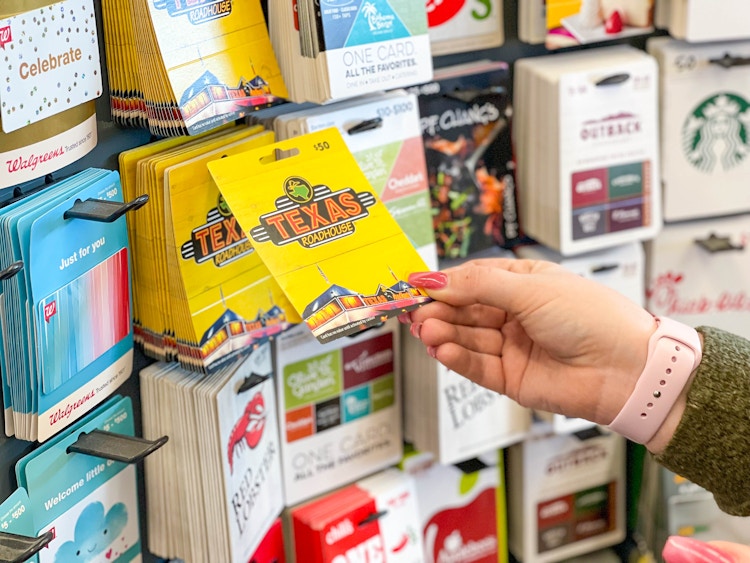 A person's hand taking a Texas Roadhouse gift card from a wall where various gift cards are hanging inside a store.