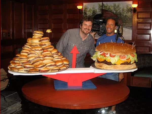 Two people standing behind a table with a large scale with an enormous burger on one side of the scale and a pile of smaller burgers on the other side.