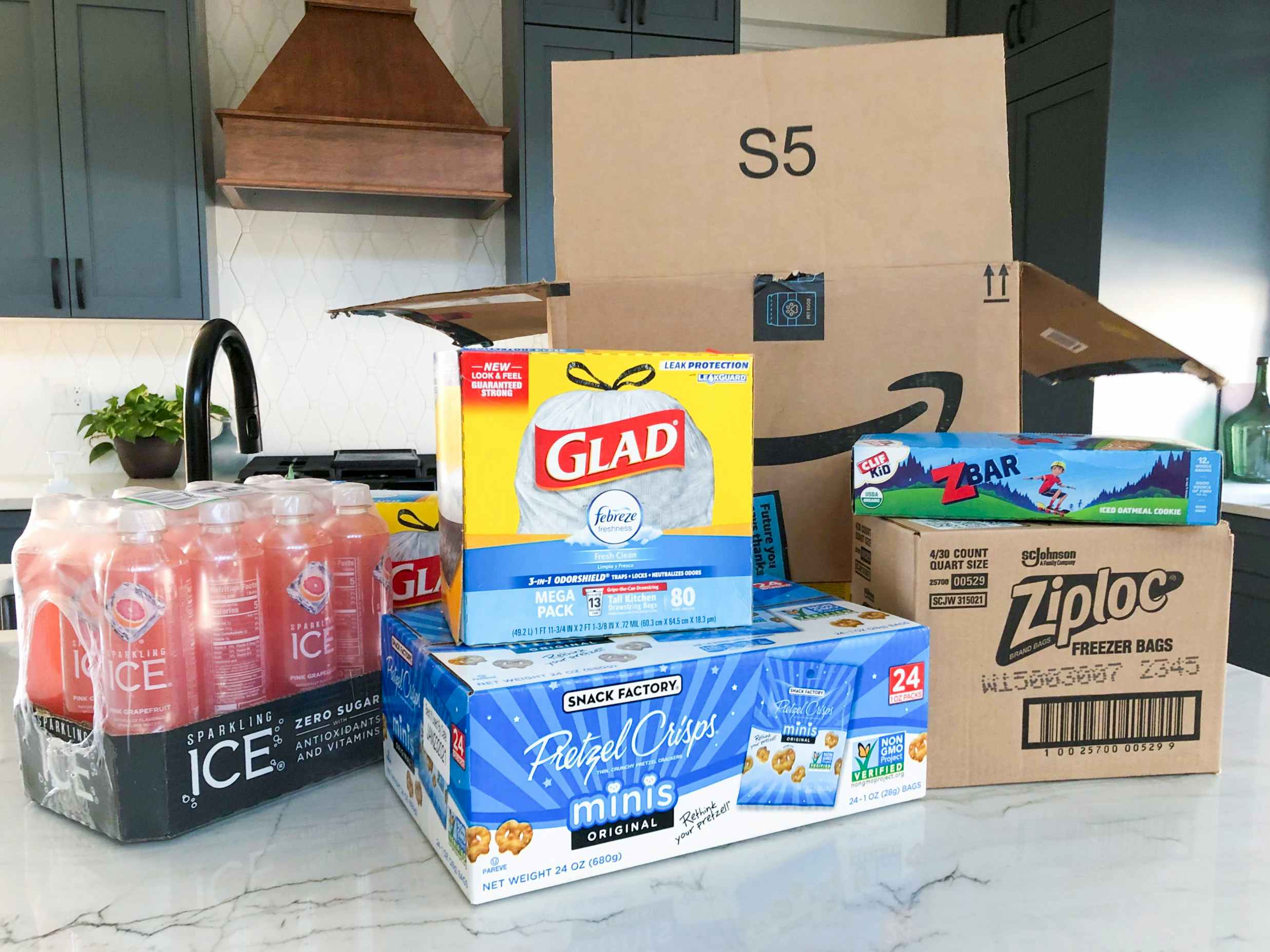 A box of Glad garbage bags, a case of ICE sparkling drinks, a box of pretzel snack bags, a box of ziplock bags, and a box of Zbars sitting on a counter in front of an Amazon Prime box.