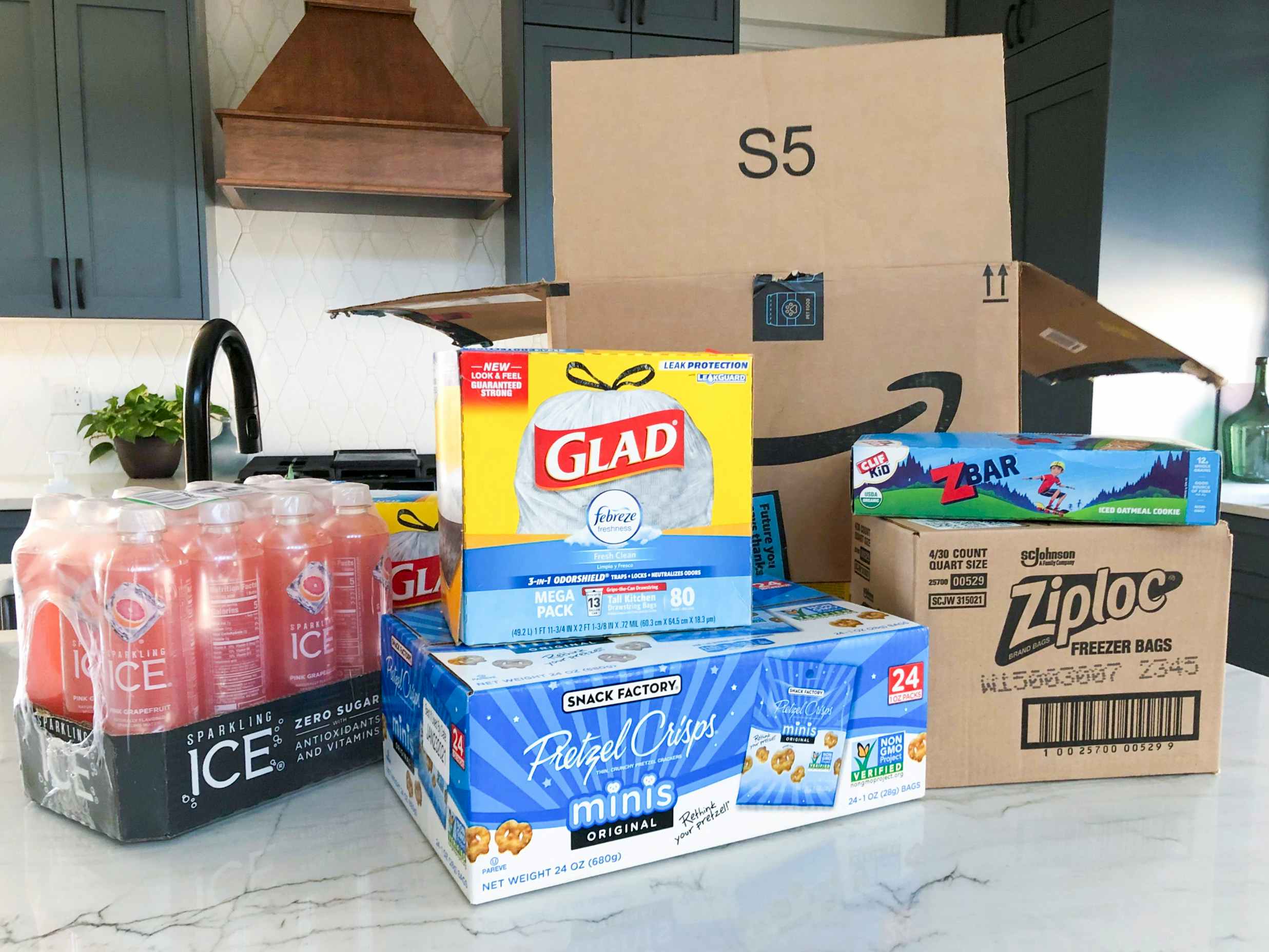 Glad garbage bags, sparking juice, pretzel snack bags, ziploc bags, and zbars sitting on a counter in front of an Amazon box