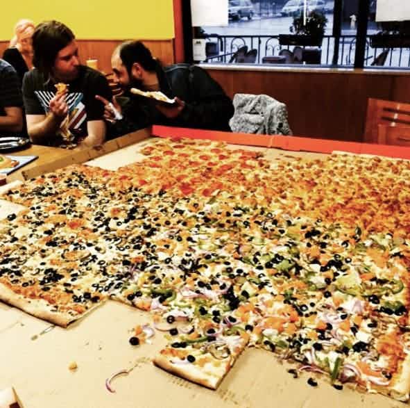 A 54-inch by 54-inch pizza sitting on a table in front of two people eating pizza.