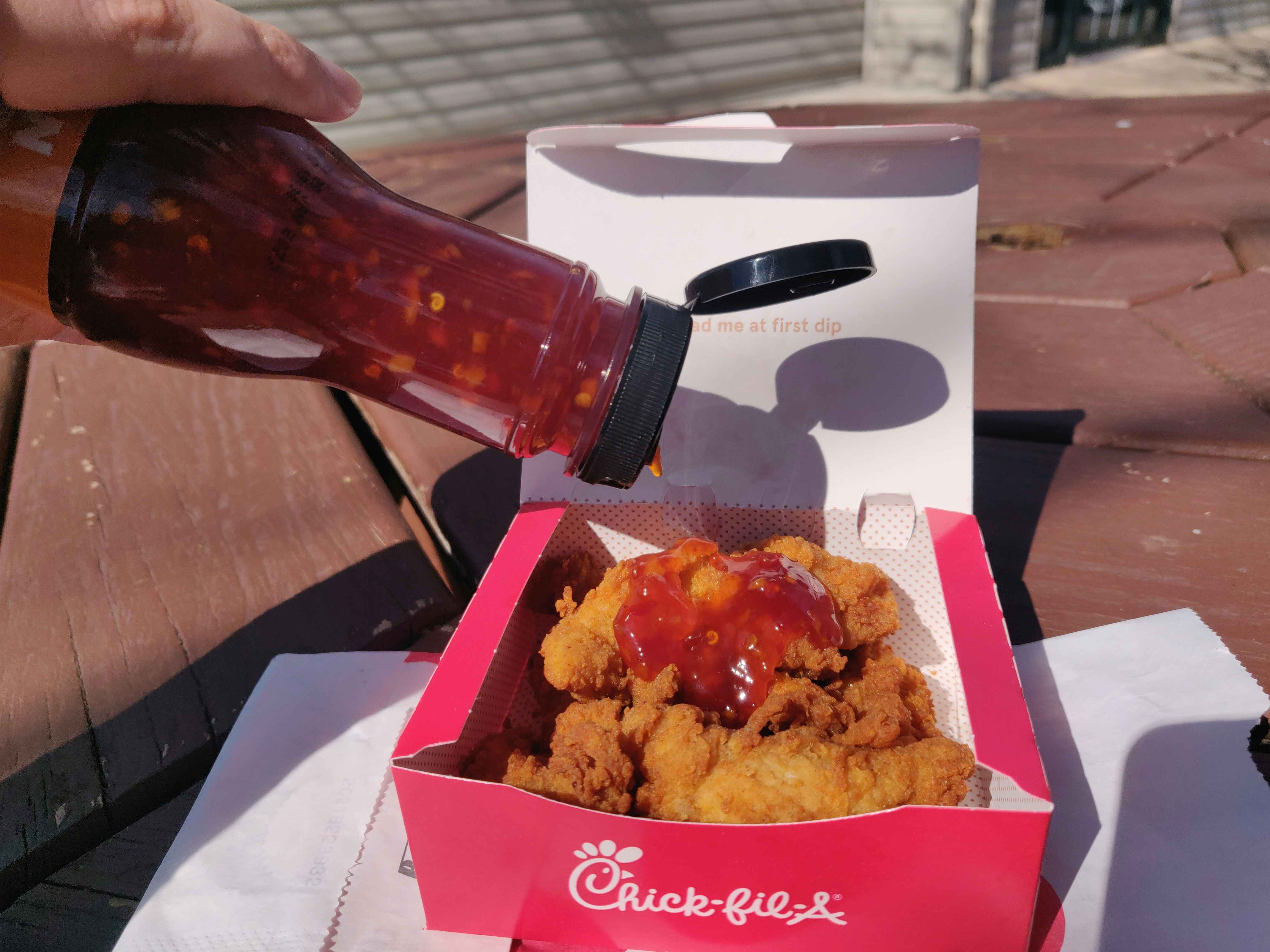 A person putting Buffalo Wild Wings sauce on top of some chicken tenders in a Chick-fil-A box.