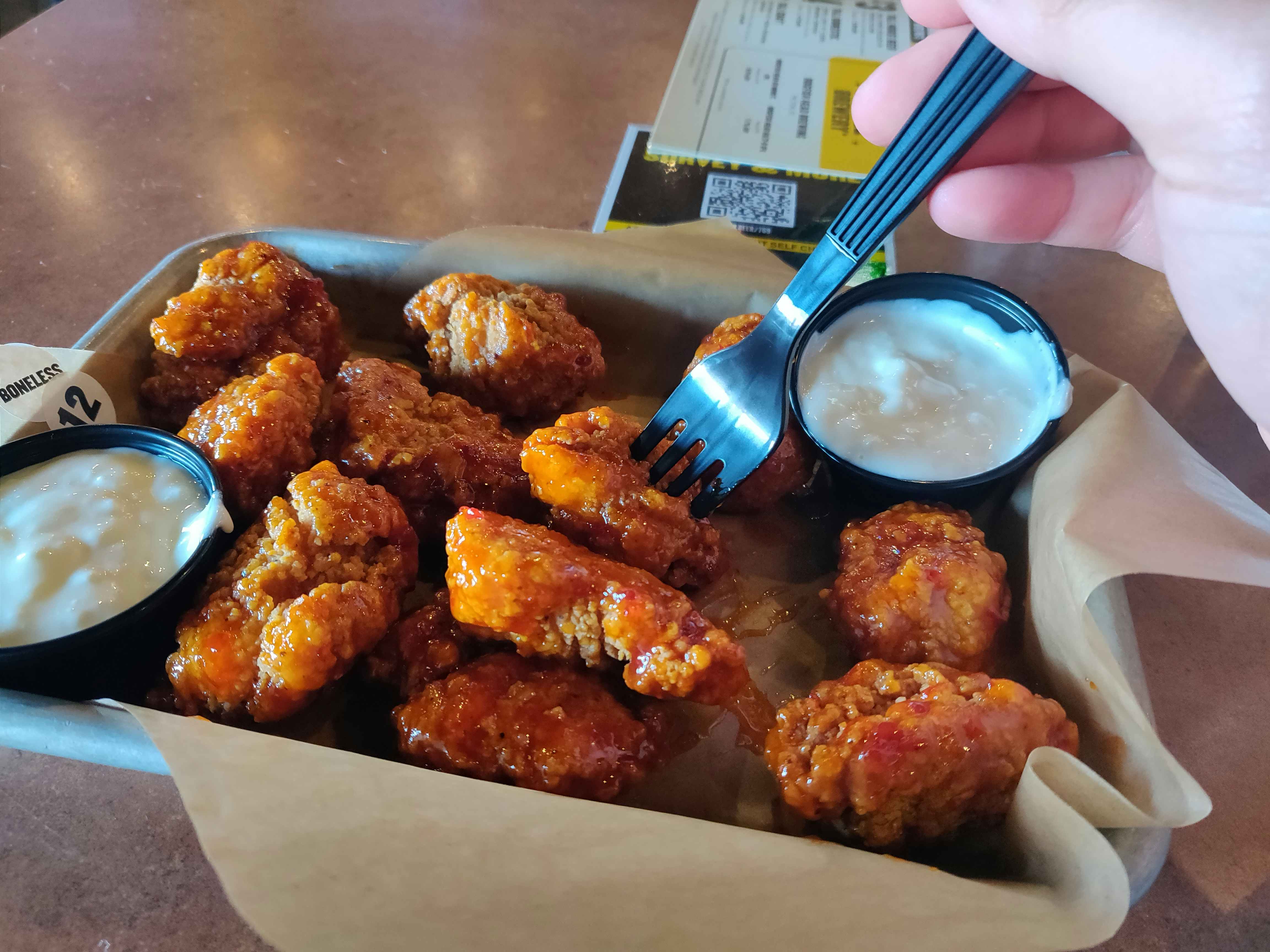 A person using a fork to pick up a boneless wing out of a platter of boneless wings and two dip cups.