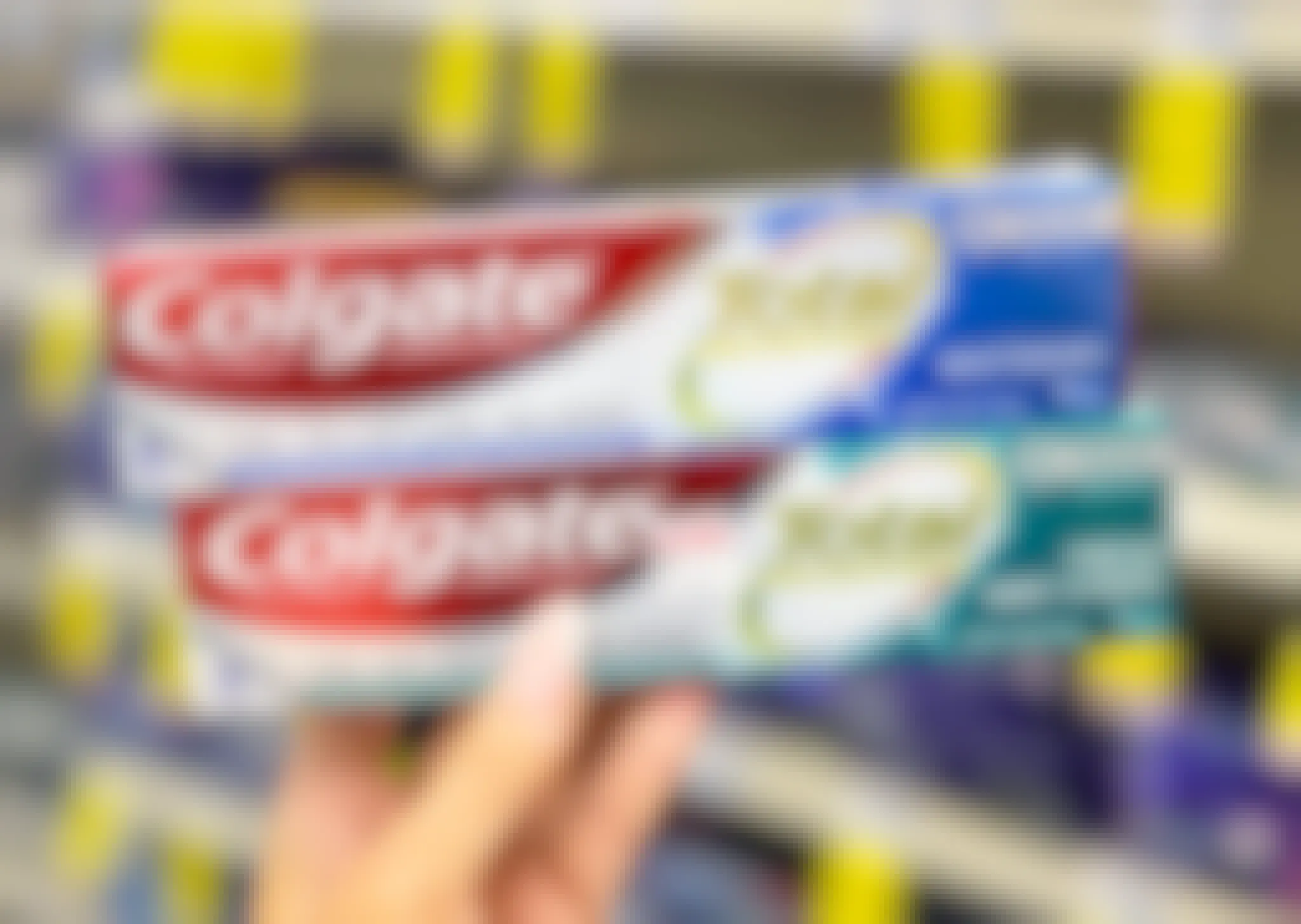 A person's hand holding up two stacked boxes of Colgate Total toothpaste. One is Whitening and the other is Fresh Mint Stripe.