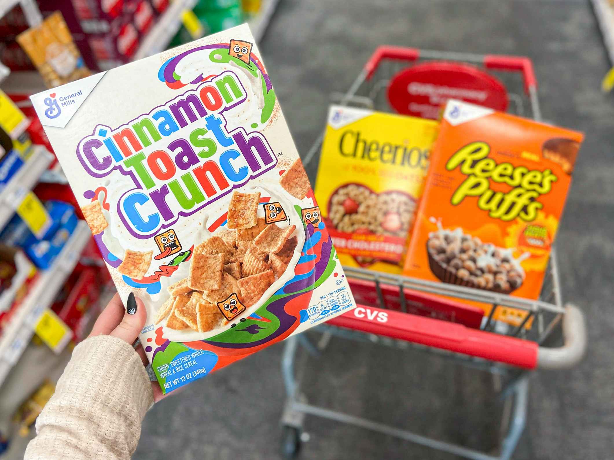 A person's hand holding a box of Cinnamon Toast Crunch in front of a box of Cheerios and a box of Reese's Puffs that are sitting in a CVS shopping cart parked in the aisle at CVS.
