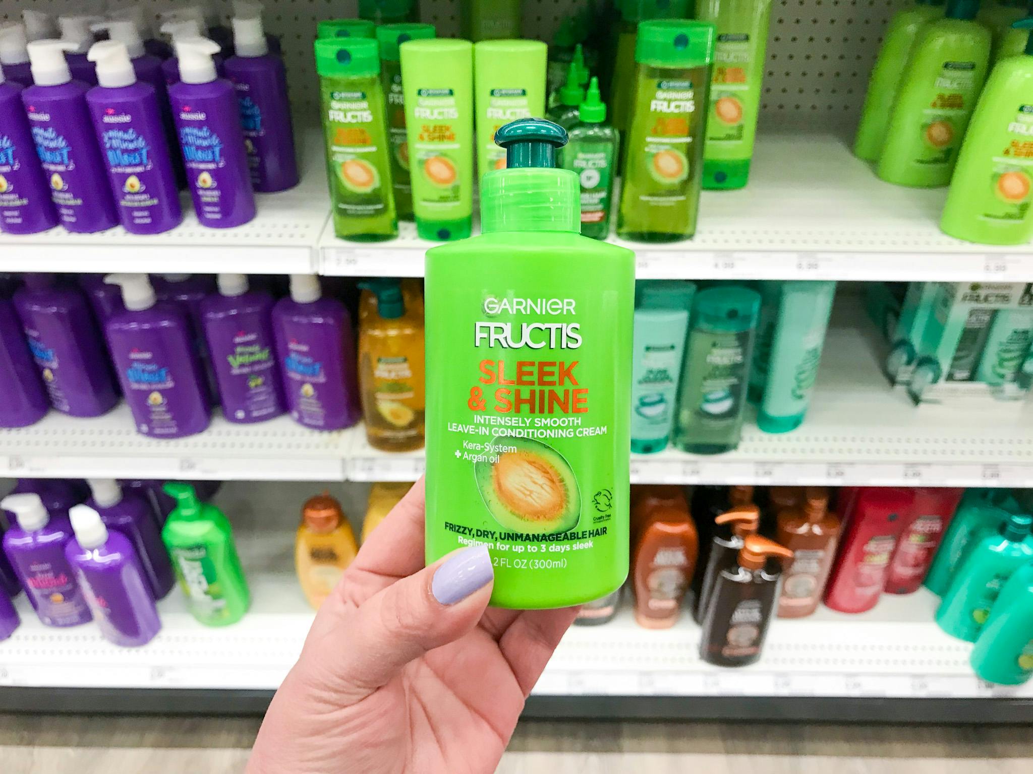 A person's hand holding up a bottle of Garnier Fructis Sleek & Shine Leave-in Conditioner in front of a shelf of haircare products at Target.